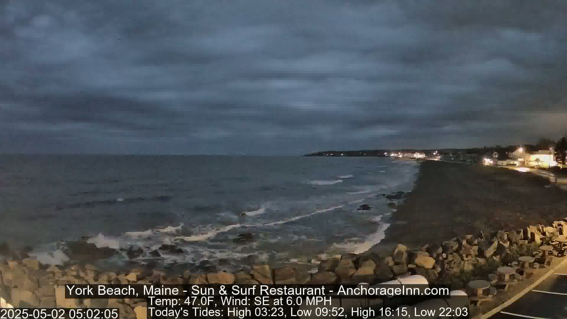 A dark, overcast night scene shows a rocky beach with gentle waves lapping the shore, under a partly cloudy sky.