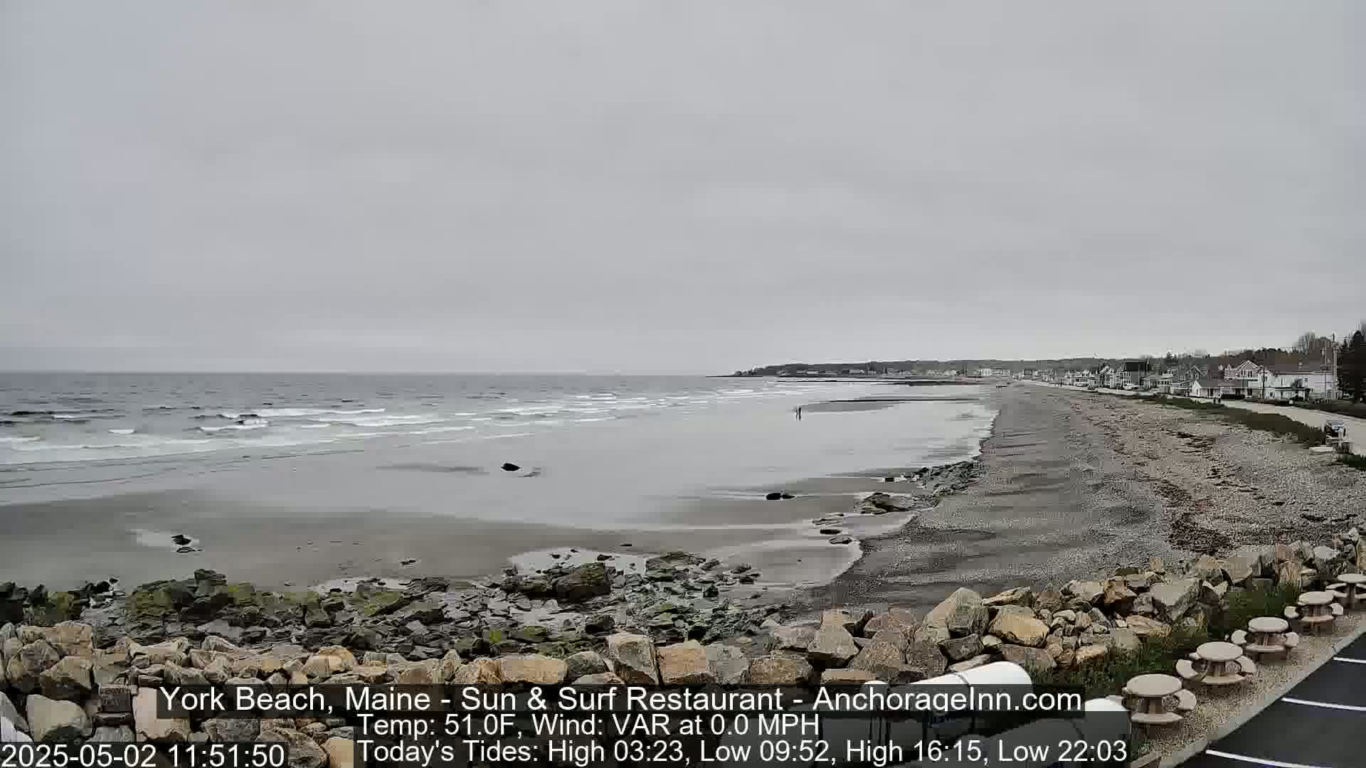 A gray, overcast day shows a long, sandy beach with small waves lapping the shore, rocky outcrops, and buildings in the distance.