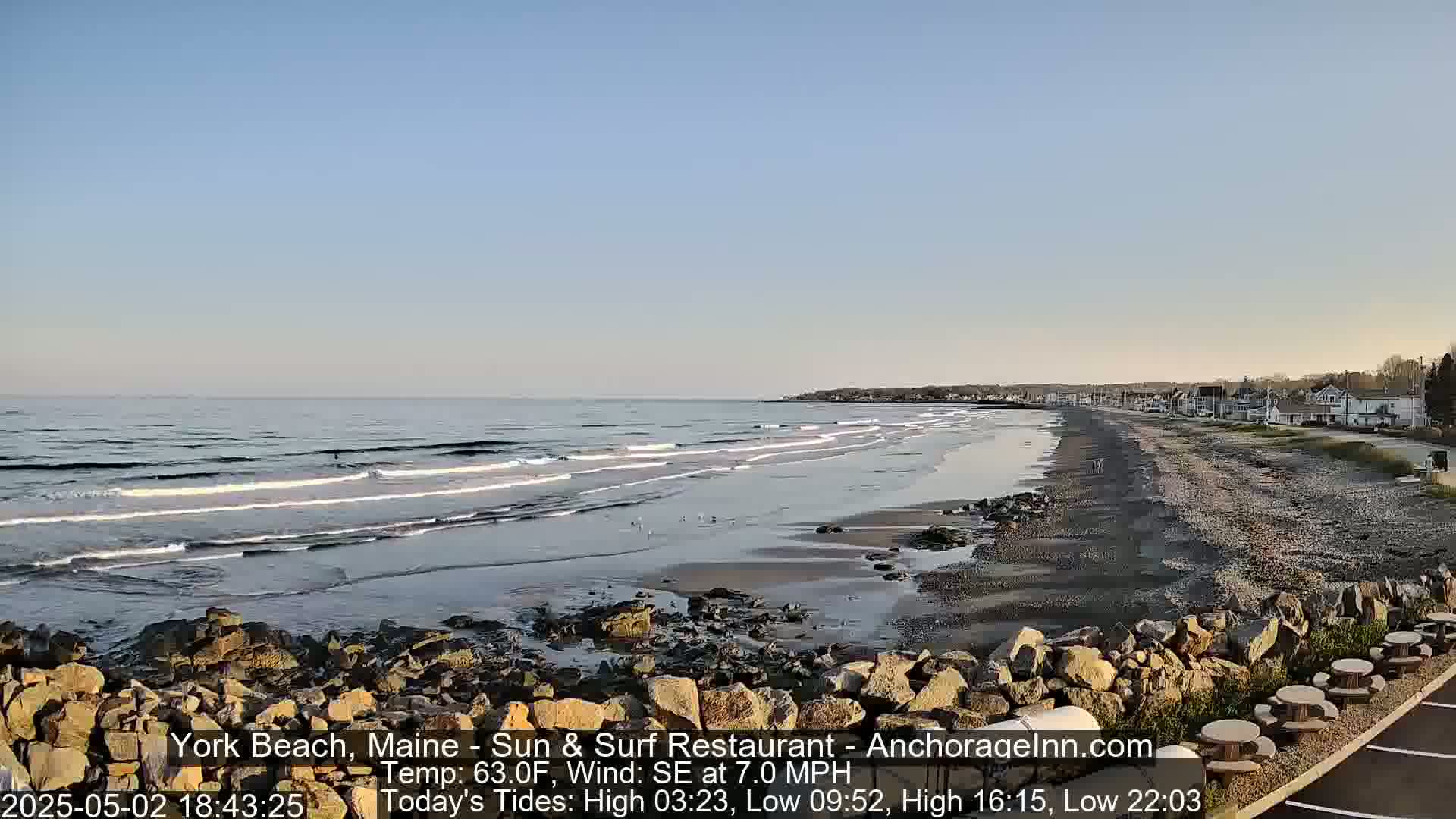 A sandy beach with gentle waves under a clear blue sky, showcasing a rocky shoreline and distant buildings.
