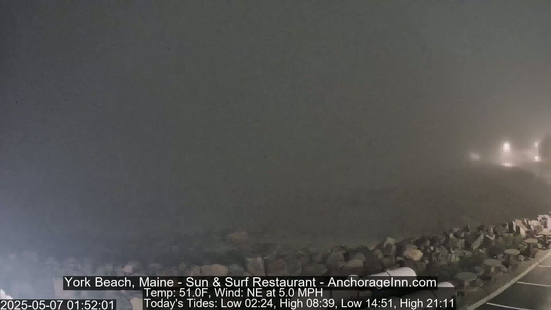 A foggy, nighttime scene shows a rocky shoreline with faint lights in the distance.