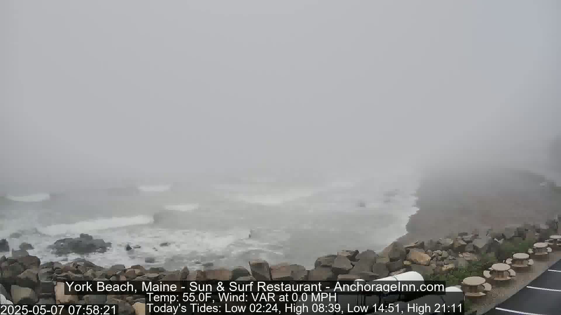 A foggy ocean scene shows waves crashing against a rocky shoreline with some outdoor seating visible in the foreground.
