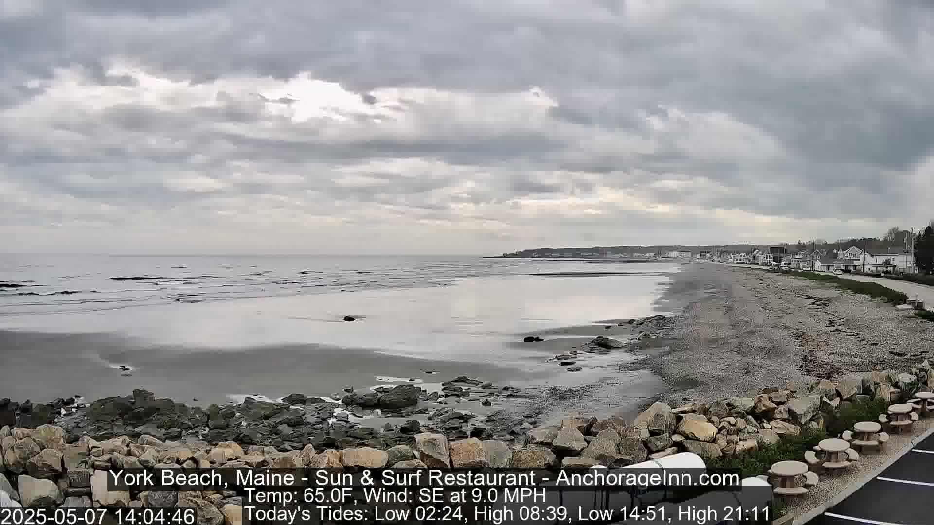 A mostly cloudy day reveals a rocky beach with a calm sea and a few buildings in the distance.