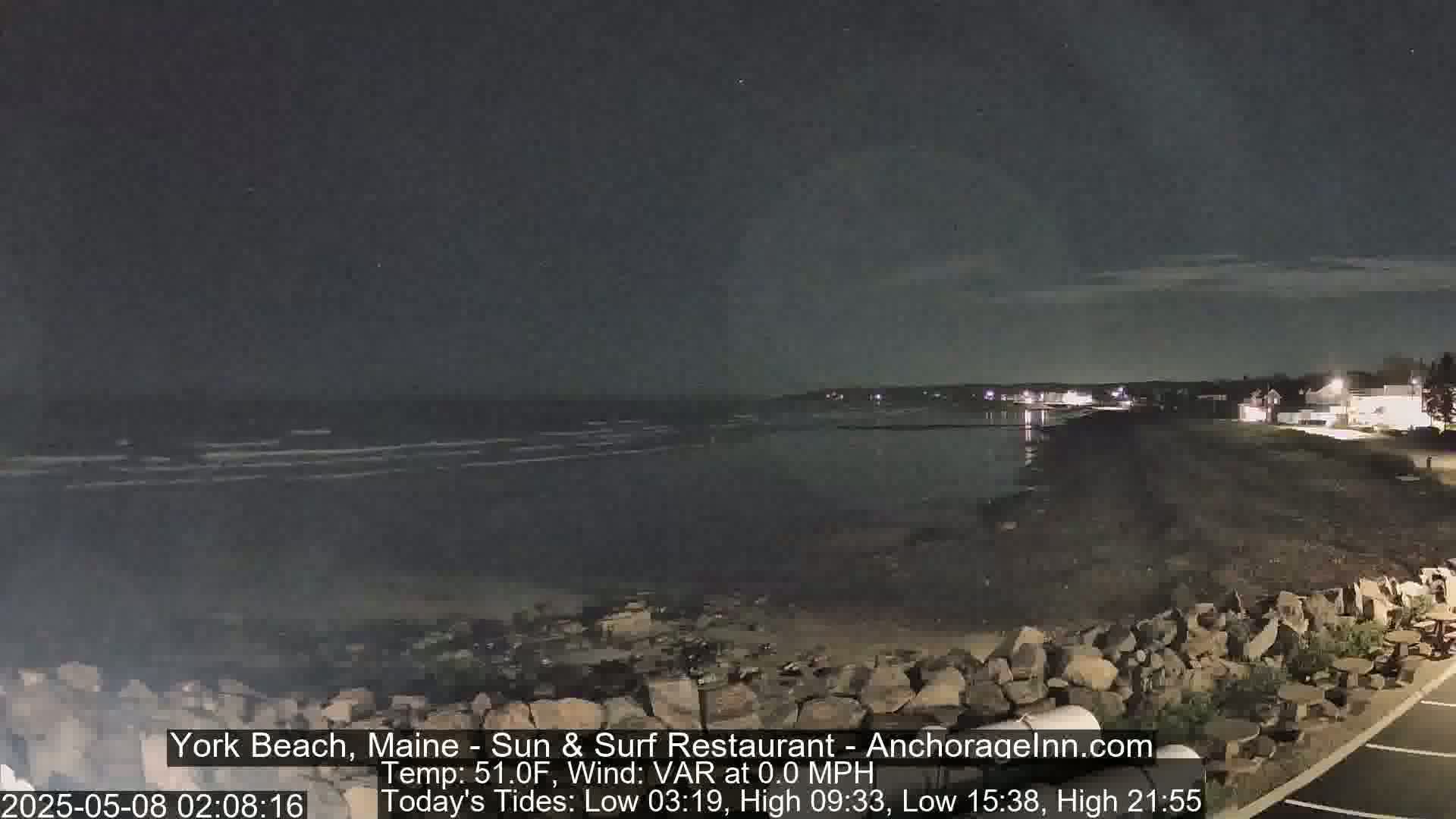 A dark, nighttime beach scene shows calm water lapping at a rocky shore under a mostly clear sky.