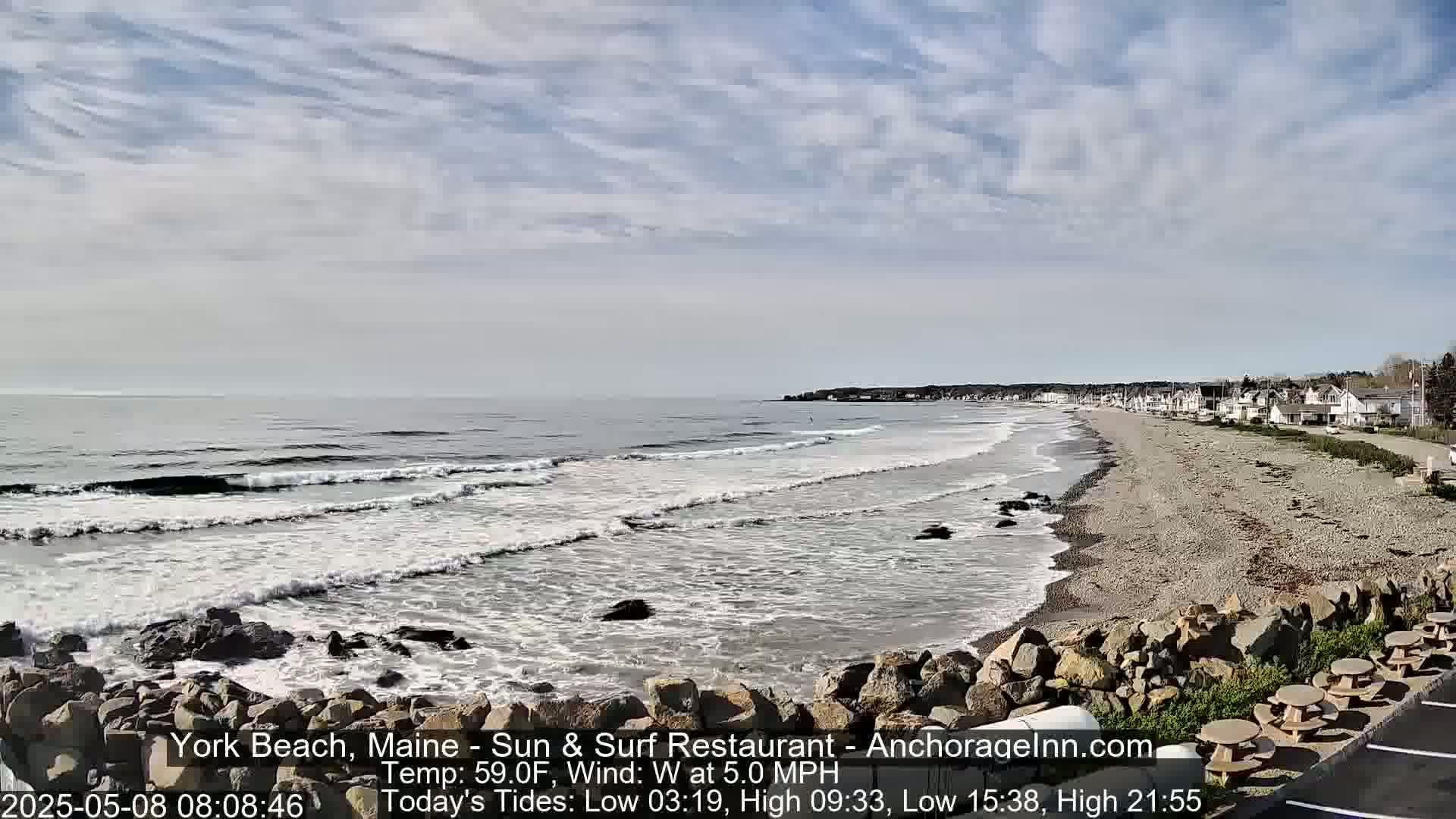 A rocky shoreline with a sandy beach and calm ocean waves under a partly cloudy sky.
