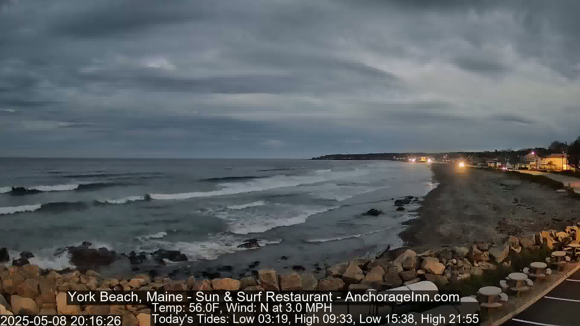A dark, overcast evening shows a rocky beach with gentle waves lapping the shore, and a few buildings visible in the distance.