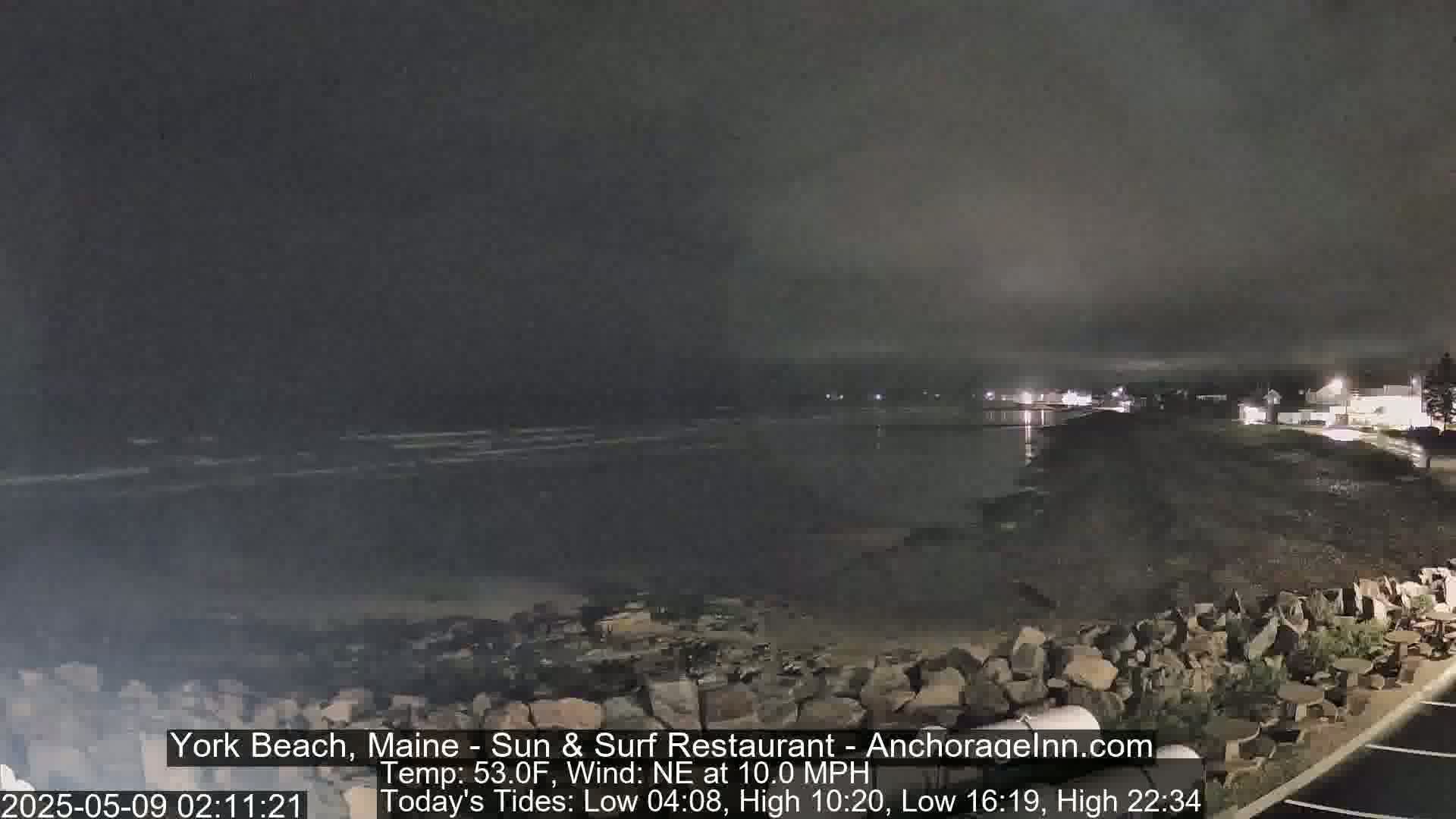 A dark, nighttime view of a rocky shoreline and calm ocean under a cloudy sky.