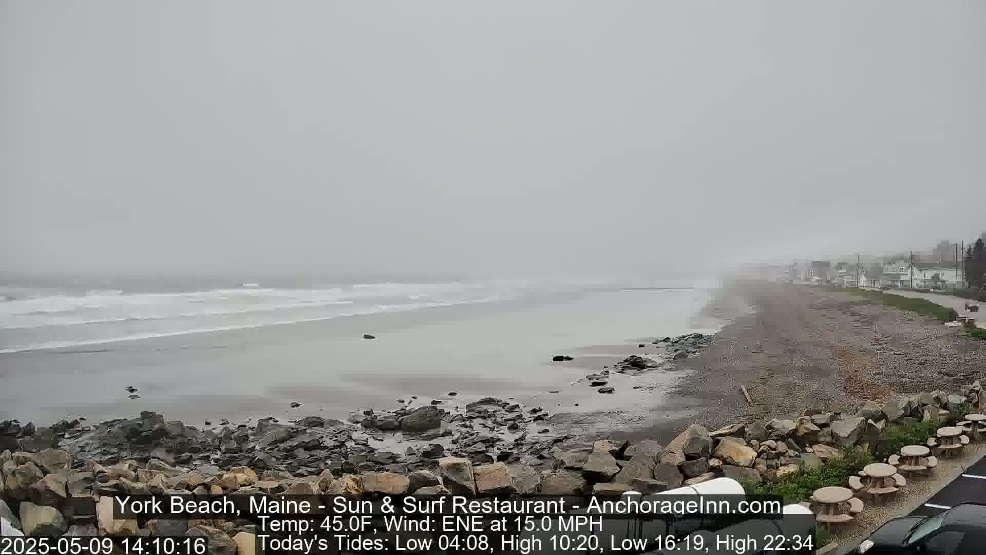 A foggy beach scene shows a rocky shoreline, gray ocean waves, and a road with buildings in the distance.