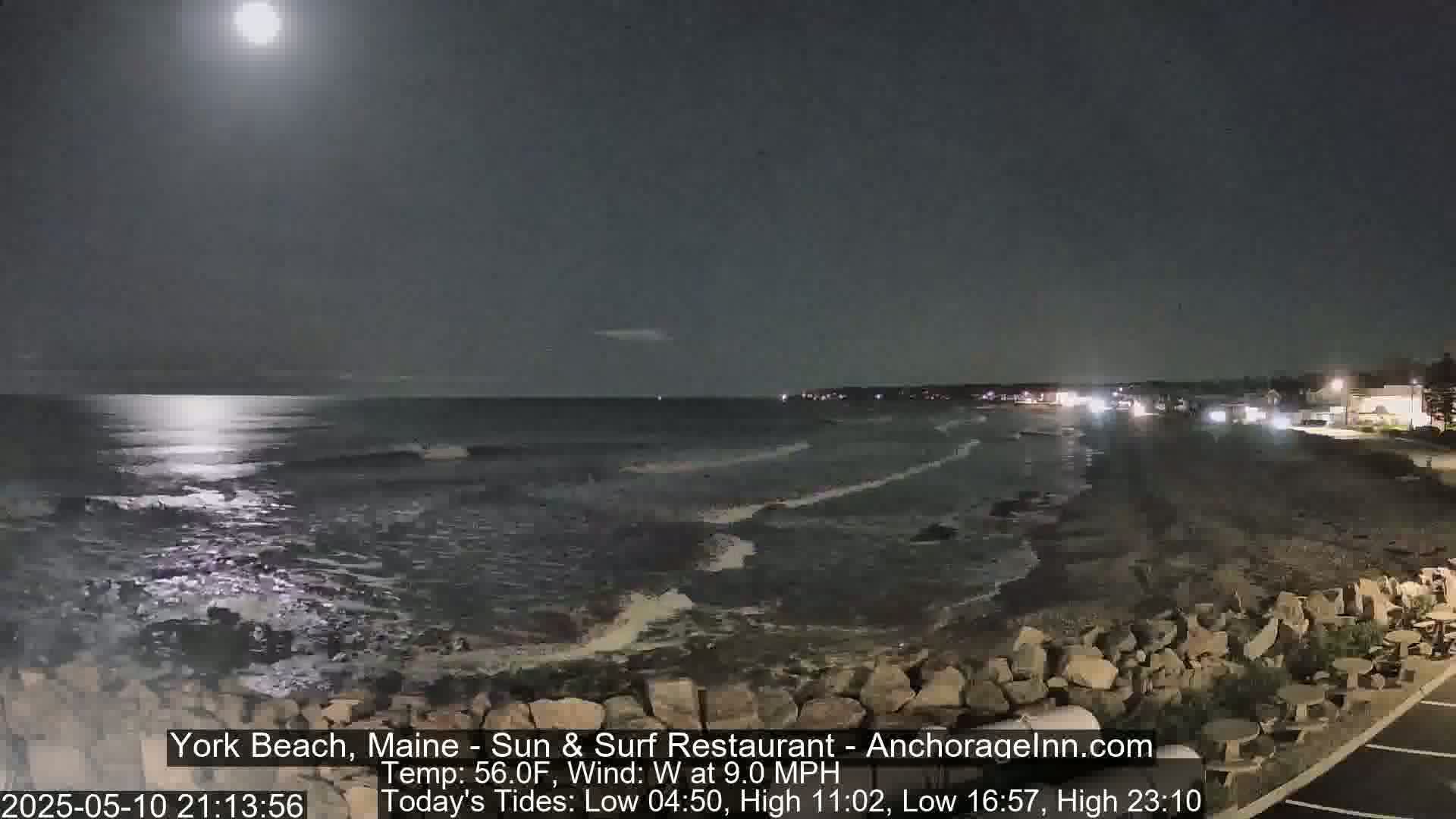 A moonlit ocean scene at night shows gentle waves lapping against a rocky shore under a partly cloudy sky with a bright moon.