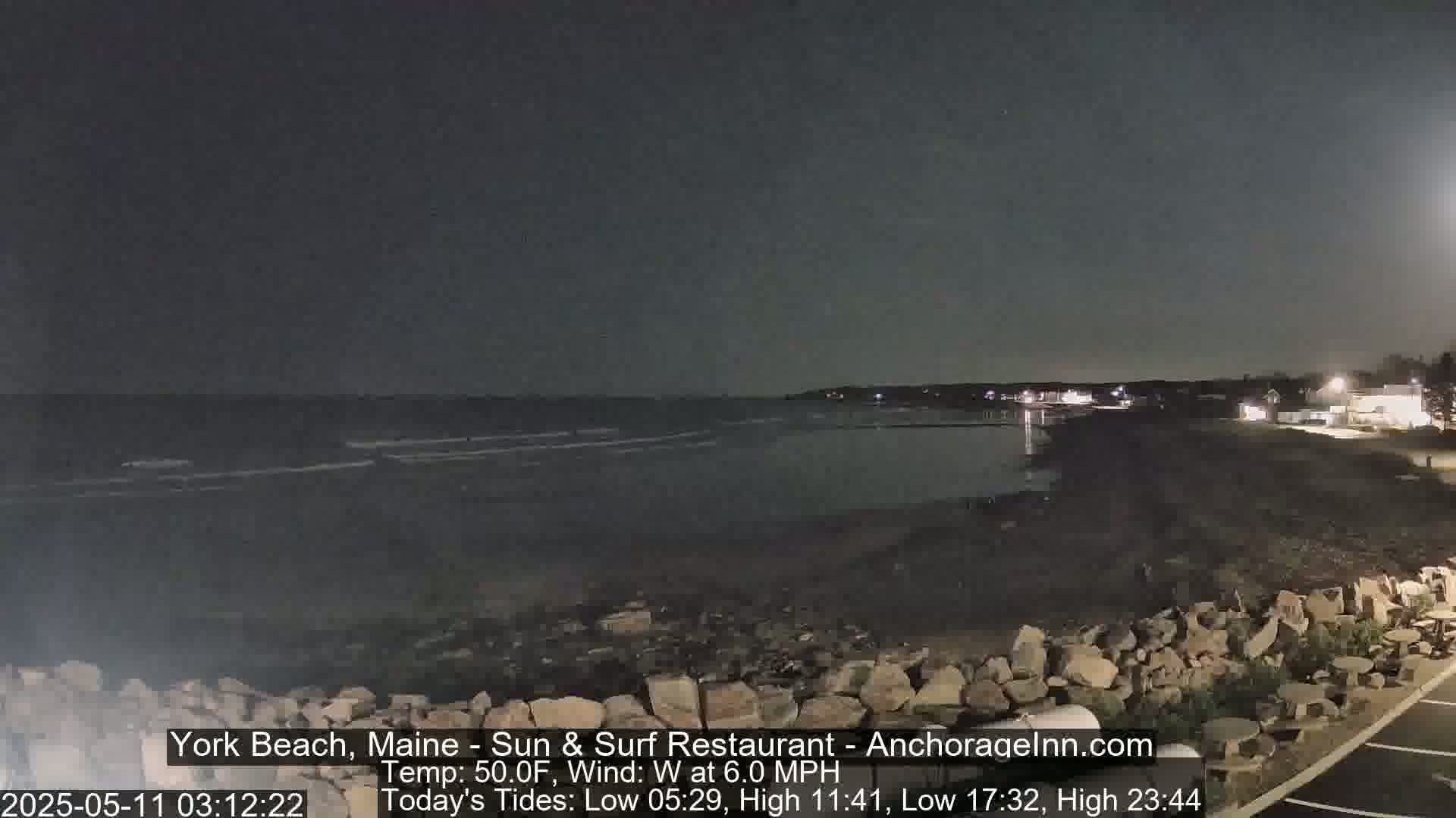 A dark nighttime beach scene shows a calm ocean under a clear, dark sky, with a rocky shoreline and distant lights.