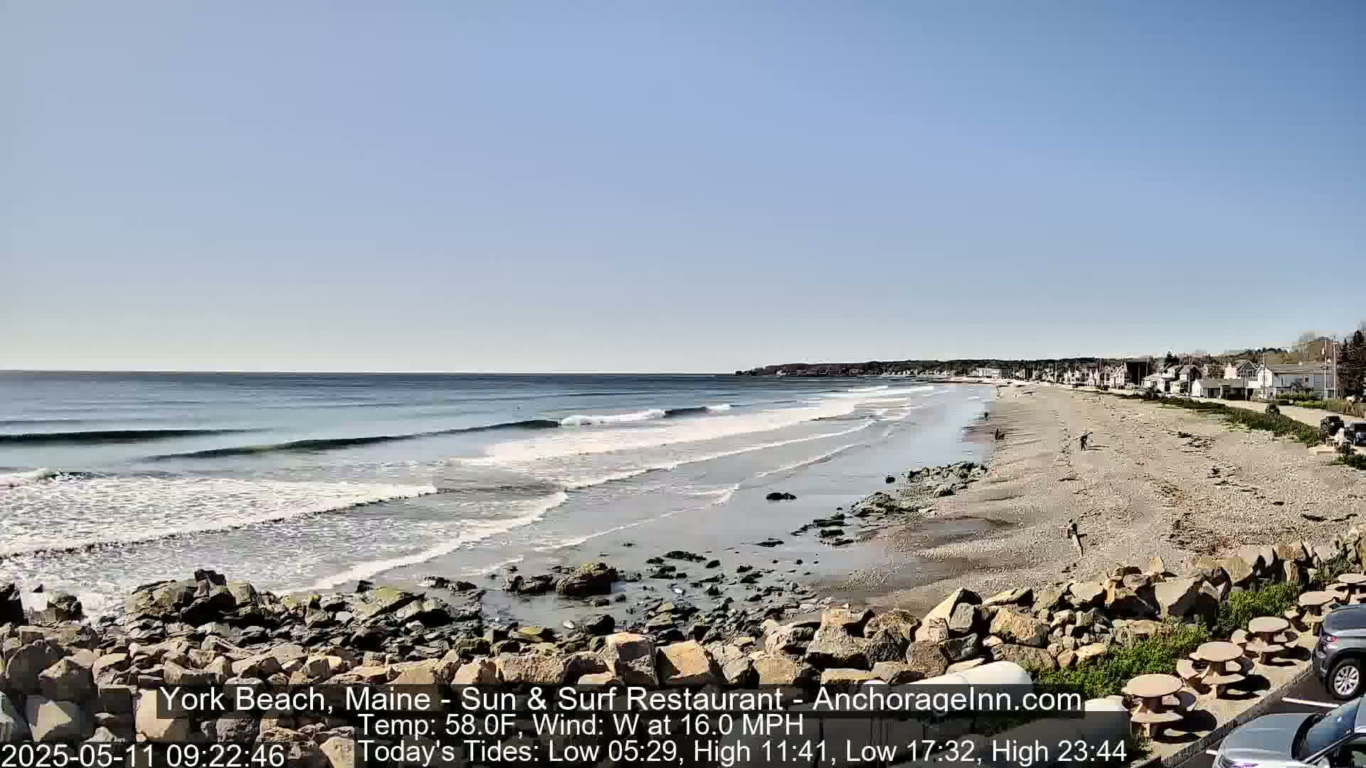 A sandy beach with small waves under a clear blue sky, bordered by a rocky shoreline and a line of houses.
