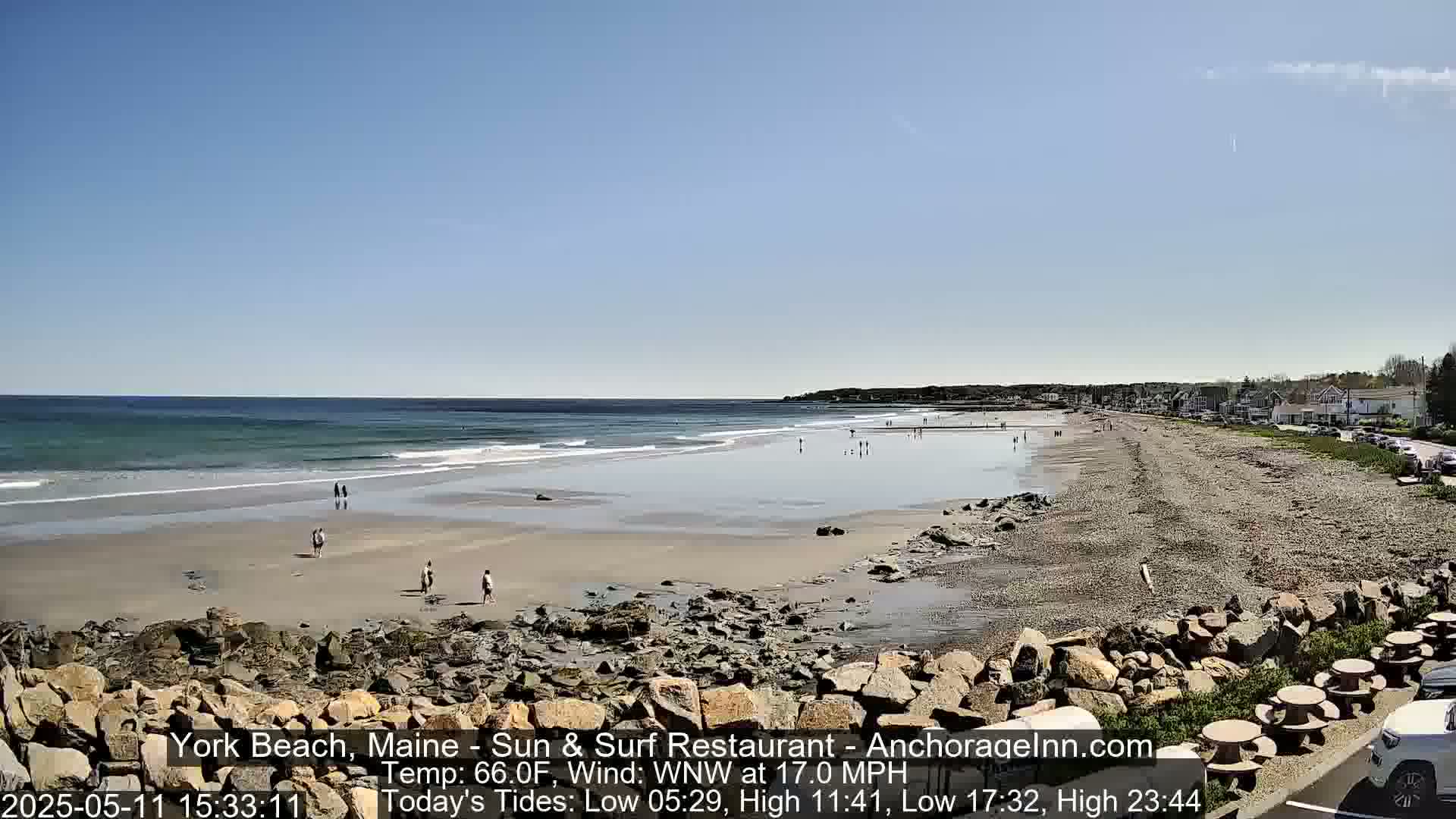 A sandy beach with a few people on it, bordered by rocks and buildings, under a clear blue sky.