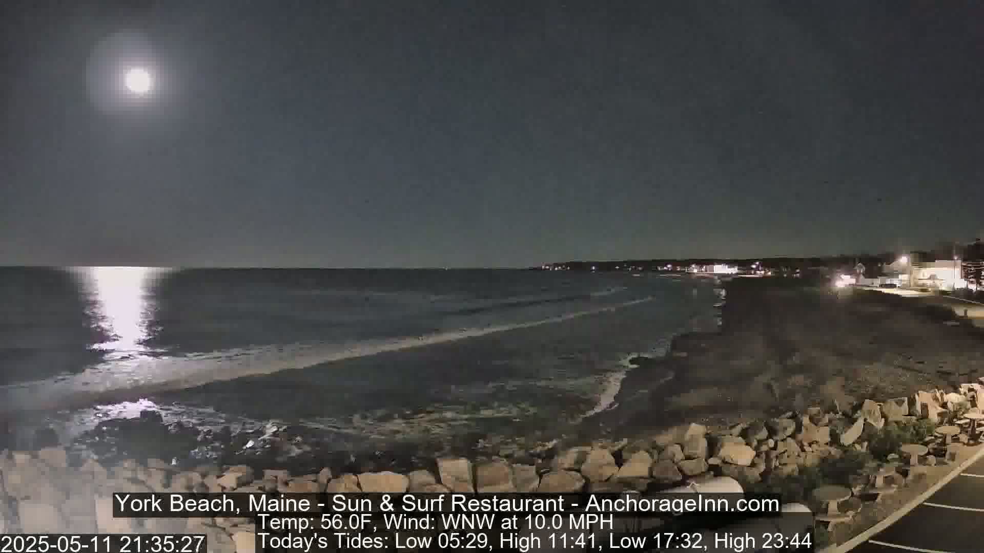 A bright moon illuminates a dark, rocky shoreline and a calm ocean under a clear night sky.