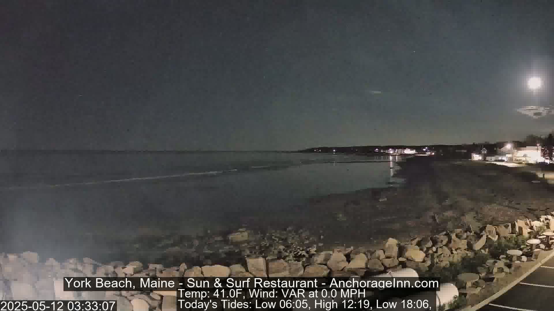 A nighttime view of a rocky beach with calm ocean waves under a bright moon.