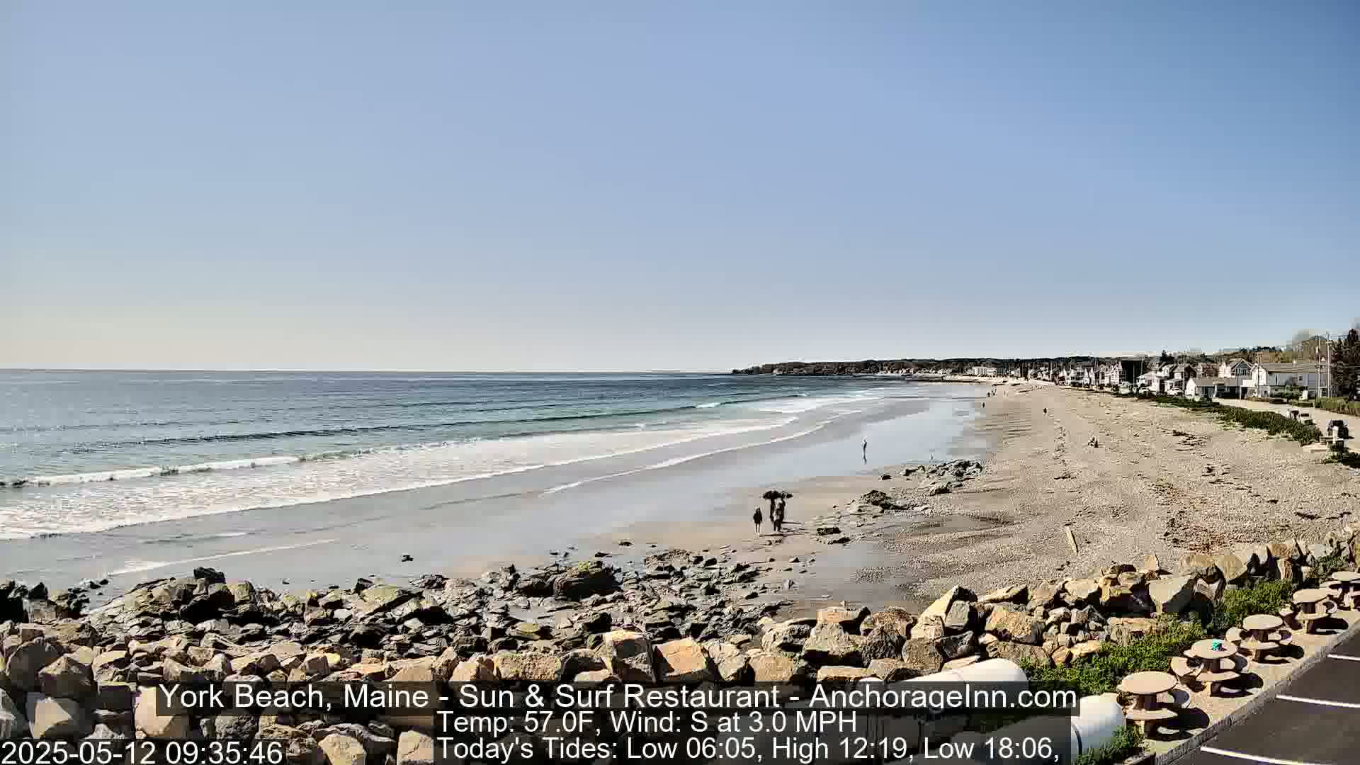 A sandy beach stretches along a calm ocean under a clear blue sky, with a few people walking on the shore and buildings visible in the distance.