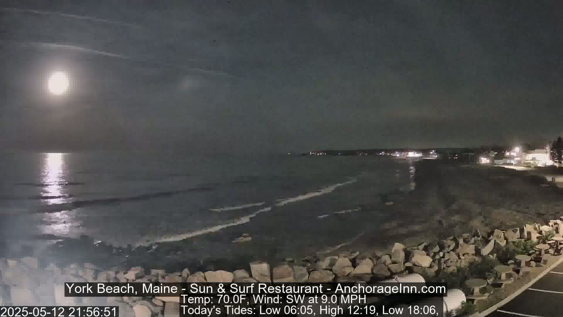 A bright moon illuminates a dark, rocky beach and calm ocean under a mostly clear night sky.
