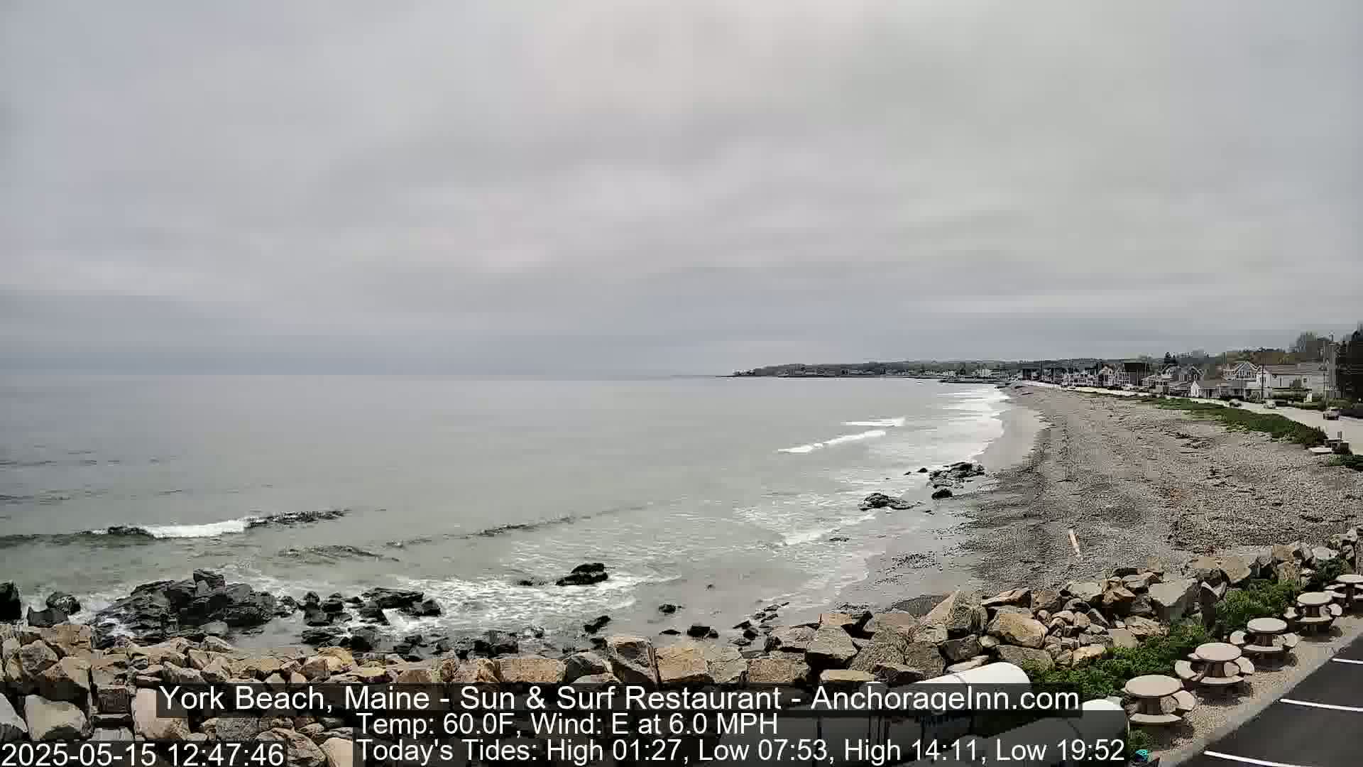 A grey, overcast day at a rocky beach with gentle waves lapping the shore, a sandy beach stretches to a line of buildings in the distance.
