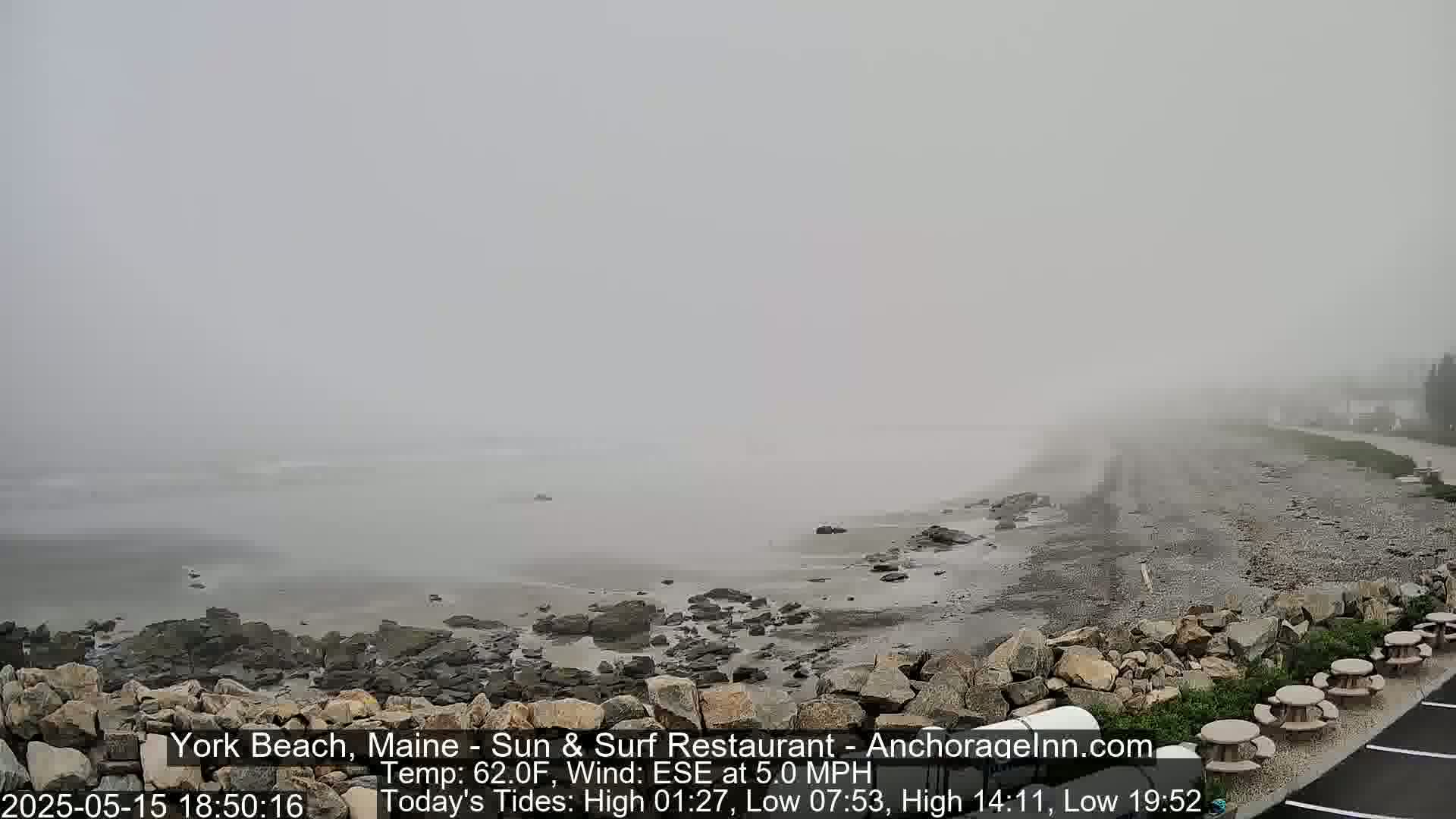 A foggy beach scene shows a rocky shoreline with a few picnic tables in the foreground, and a calm, misty ocean extending to the horizon.