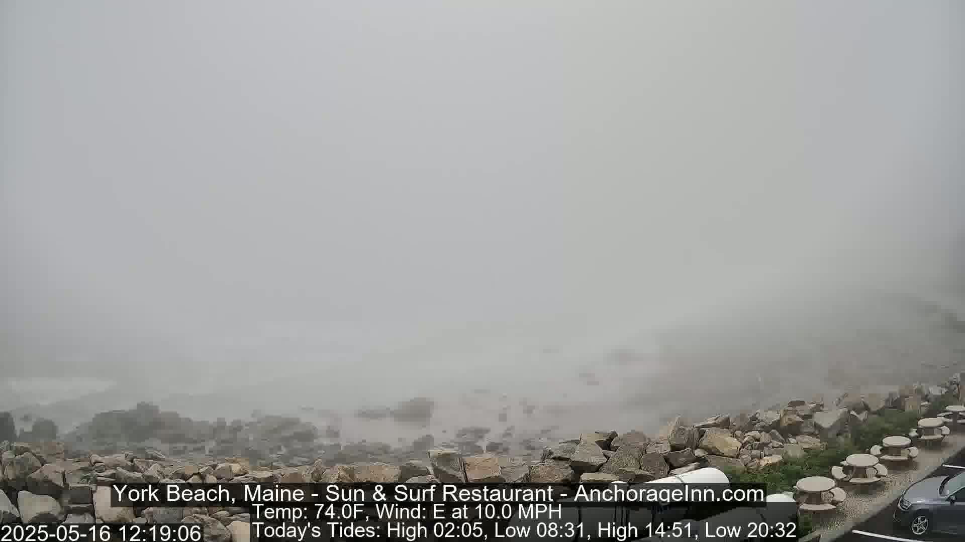 A foggy coastal scene shows a rocky shoreline with outdoor seating and a parking lot in the foreground.