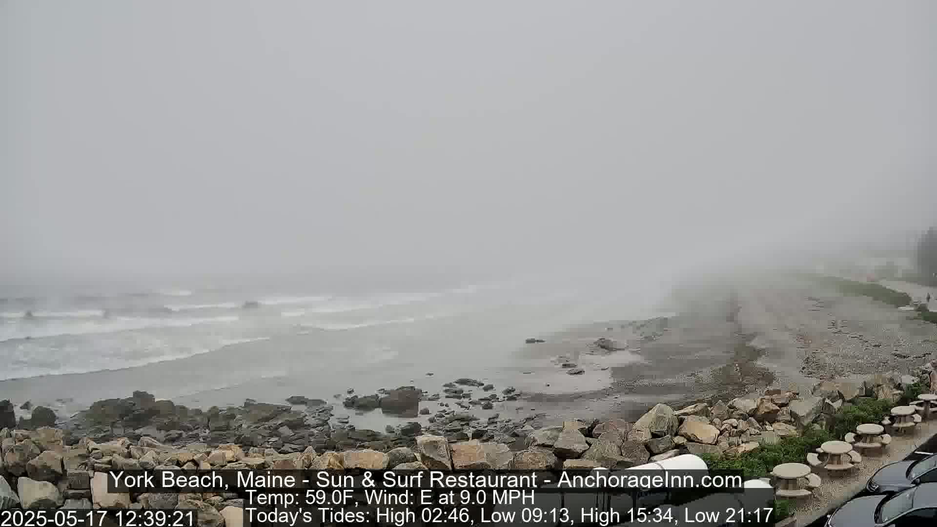 A foggy beach scene shows waves crashing against a rocky shore with some picnic tables and cars visible in the foreground.