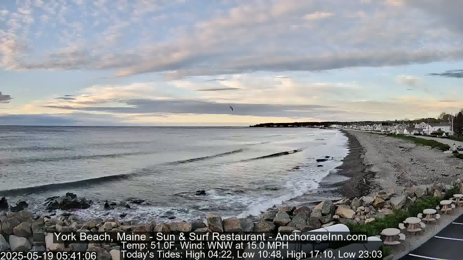 A beach with small waves rolls onto a rocky shore under a partly cloudy sky.