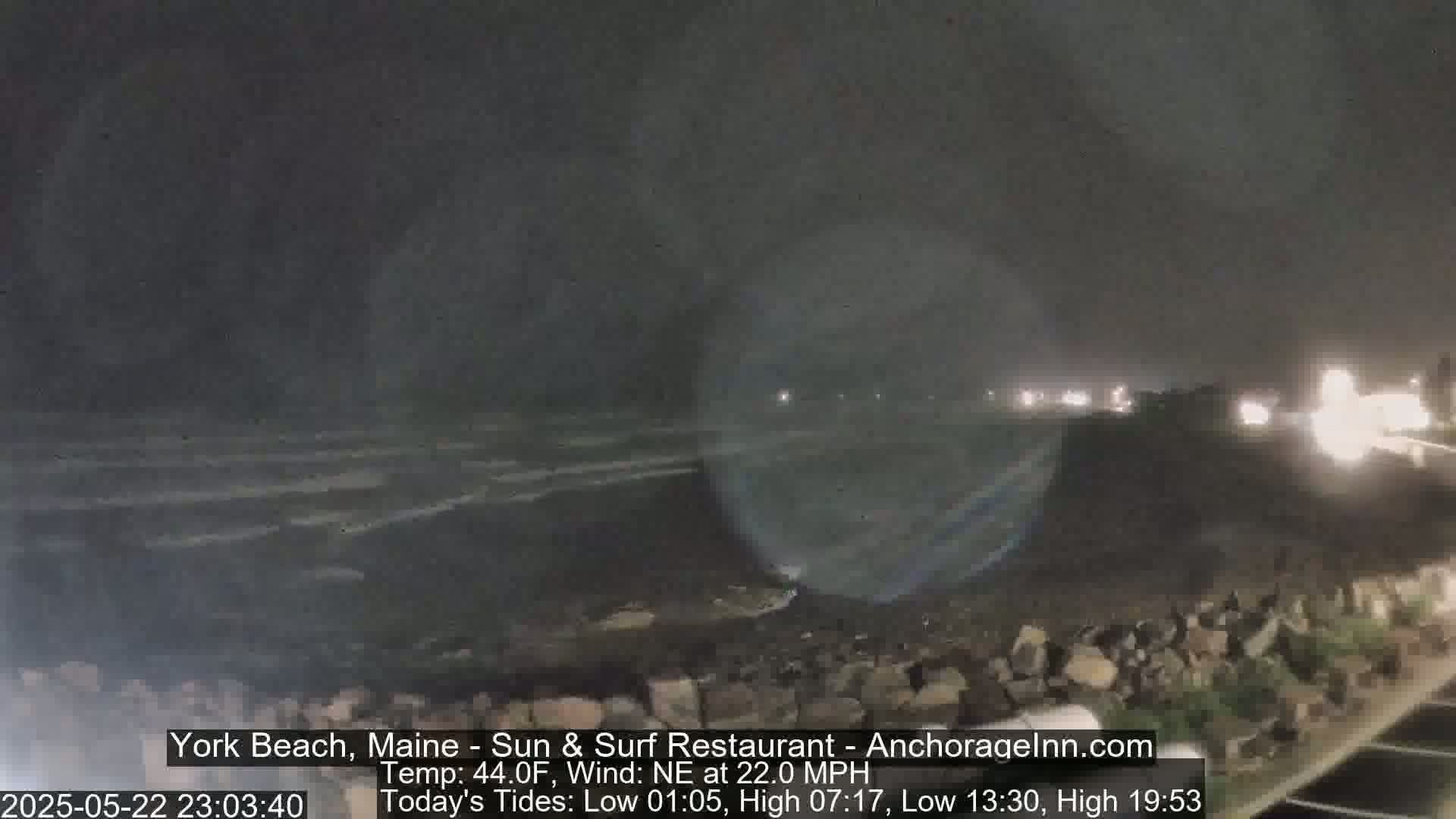 A nighttime coastal scene shows dark, choppy waves under a cloudy sky, with a rocky shoreline and distant lights visible.