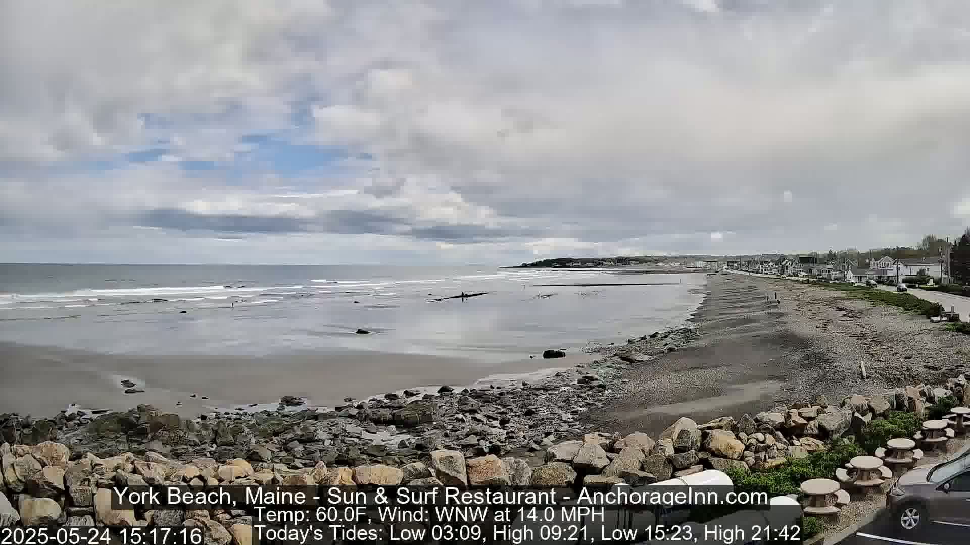 A partly cloudy day shows a rocky beach with a calm ocean and a few people visible in the distance.