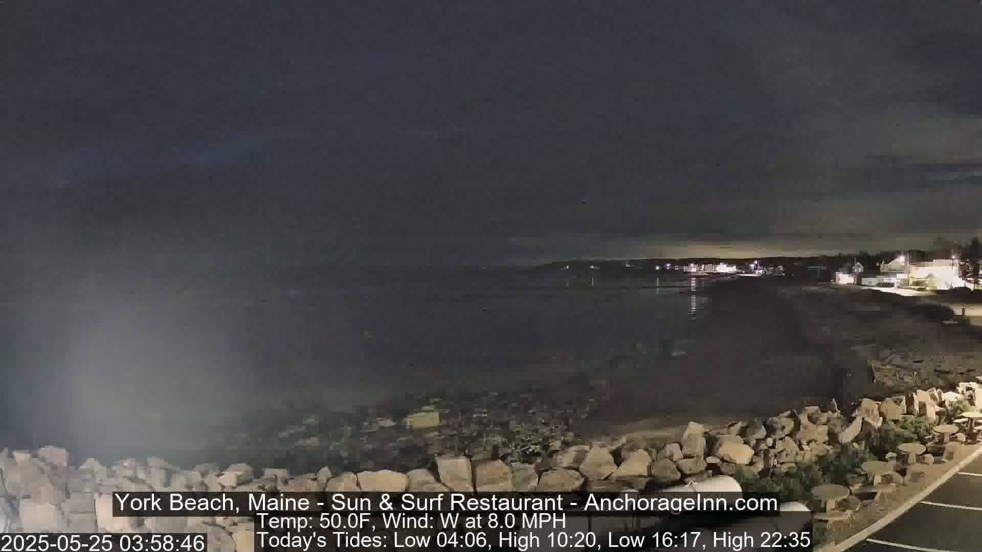 A nighttime coastal scene shows a dark body of water under a mostly clear, dark sky, with a rocky shoreline and dimly lit buildings in the distance.