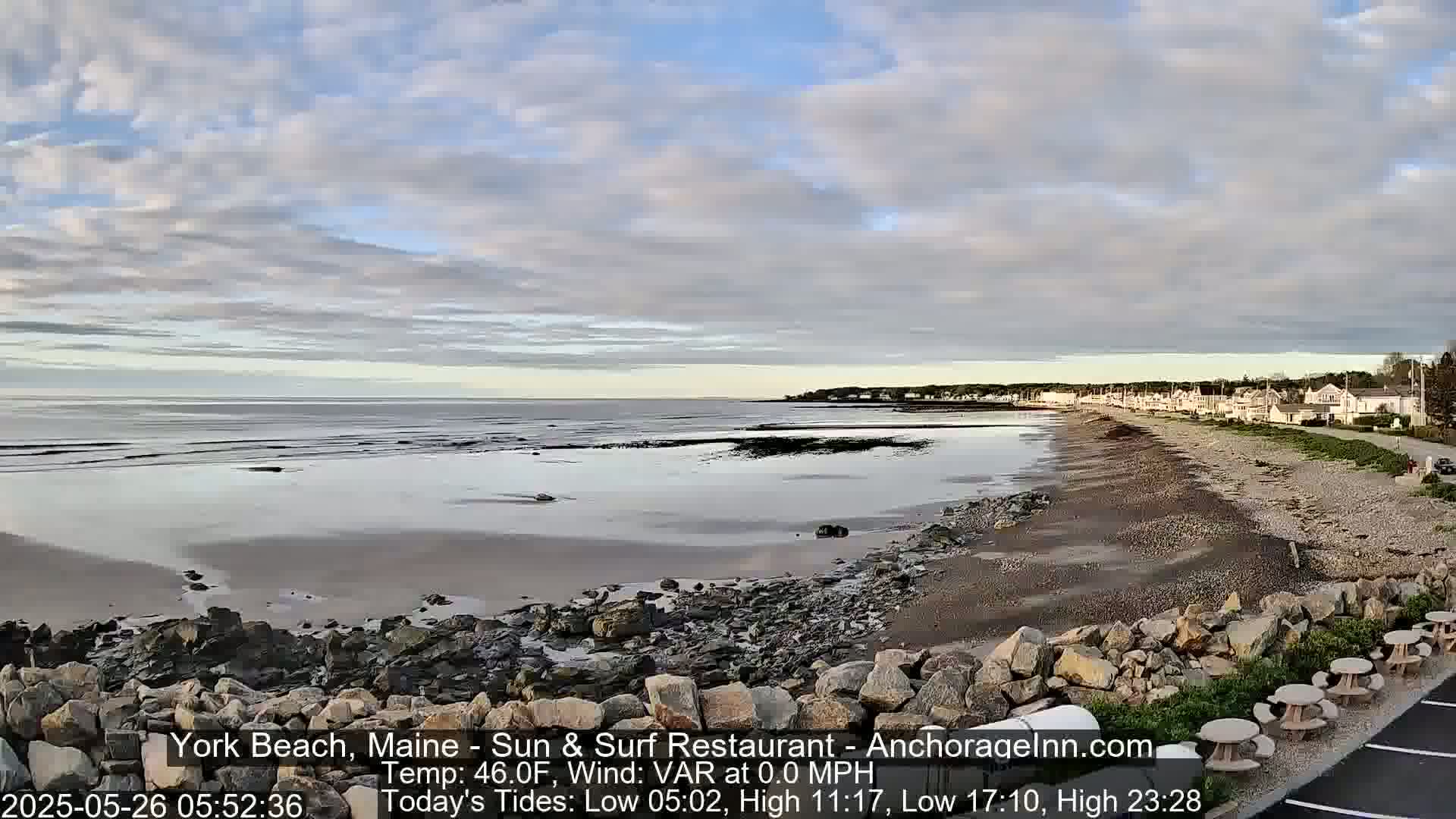 A rocky beach with calm, shallow water stretches out to a quiet seaside town under a partly cloudy sky.