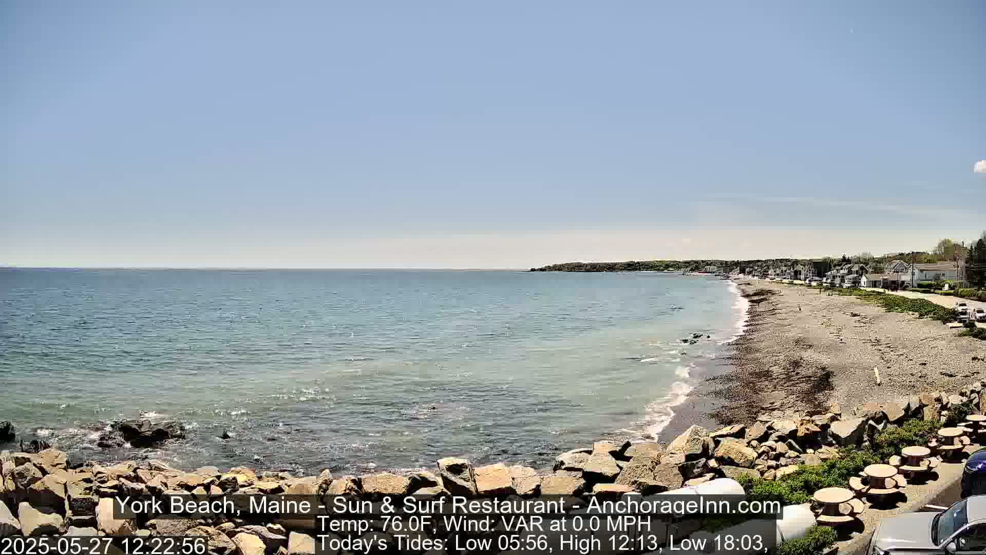 A rocky shoreline with a calm, light-blue ocean under a clear blue sky, showing a sandy beach with a few people and buildings in the distance.
