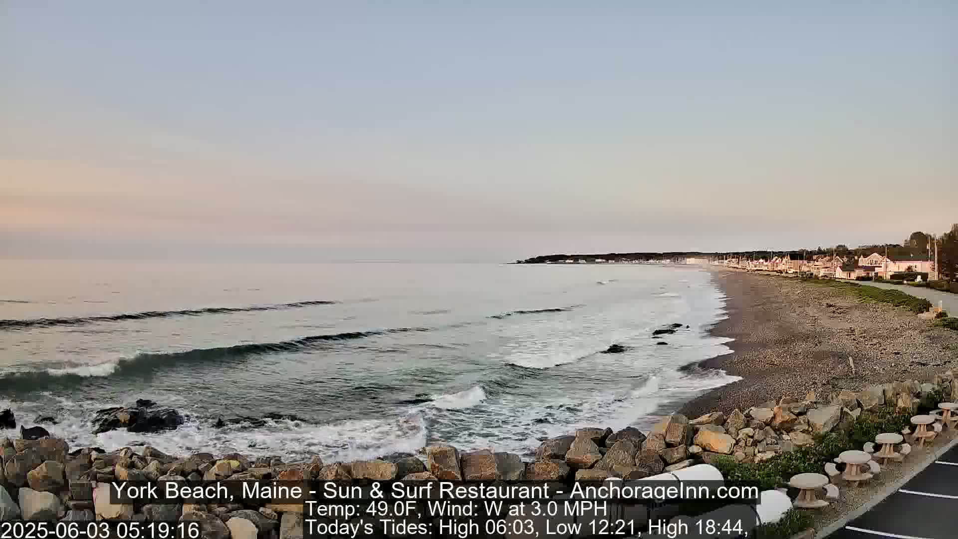 A calm ocean with small waves laps a sandy beach backed by a row of houses under a pale, clear sky.