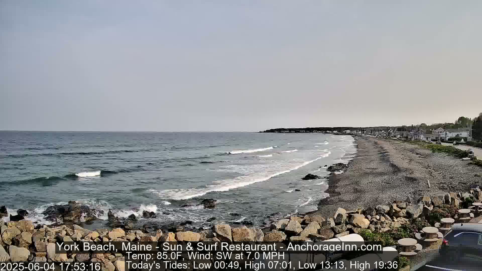 A rocky shoreline with a sandy beach and gentle waves stretches along a coast under a mostly clear sky.