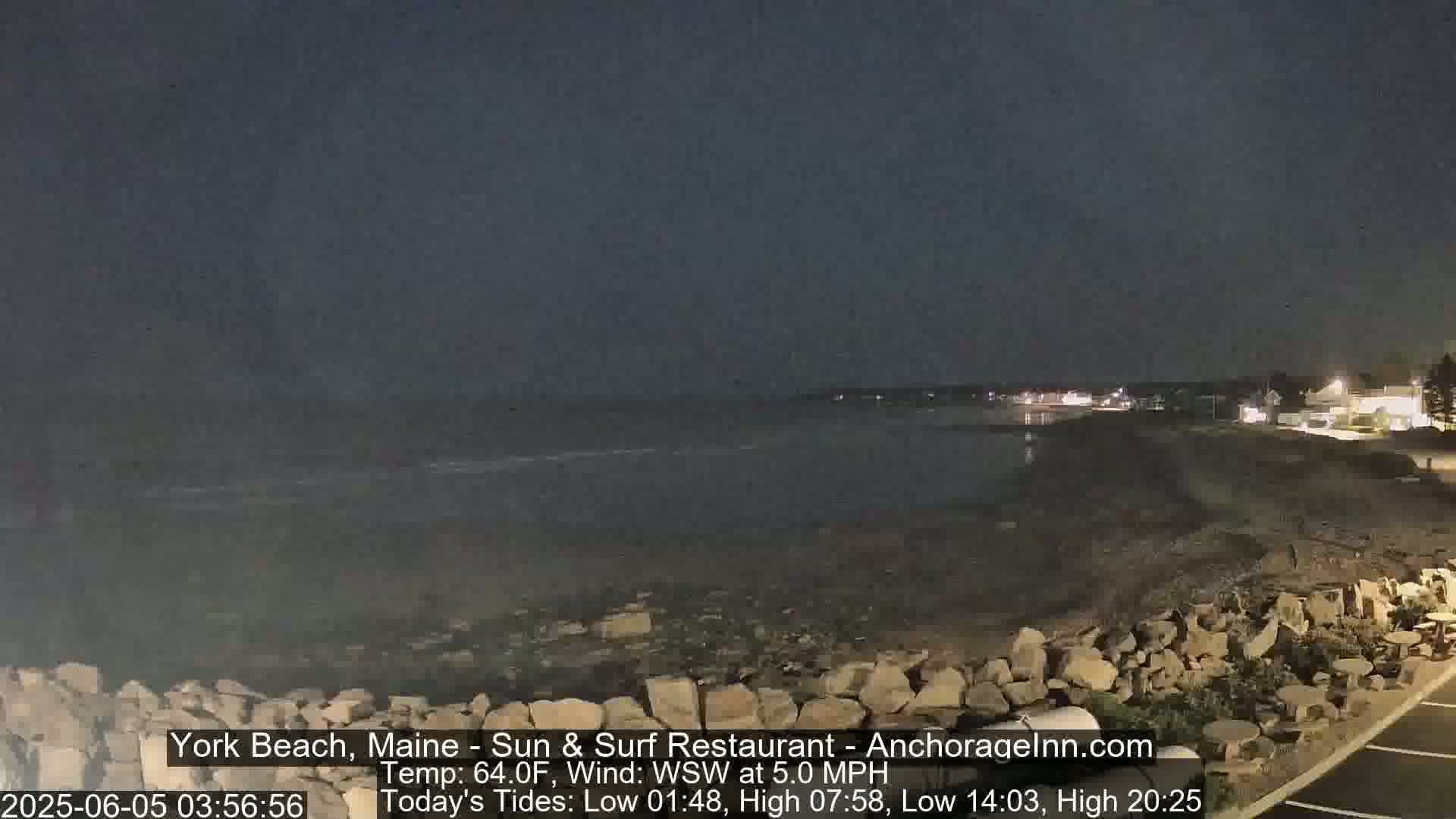 A dark, nighttime beach scene shows a rocky shoreline and calm water under a mostly clear sky.