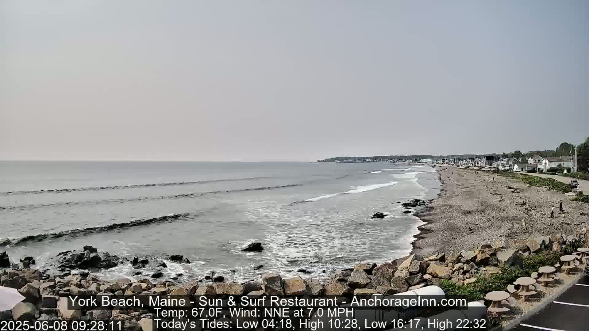 A long, sandy beach with small waves and a few people stretches out to a calm ocean under a hazy sky.