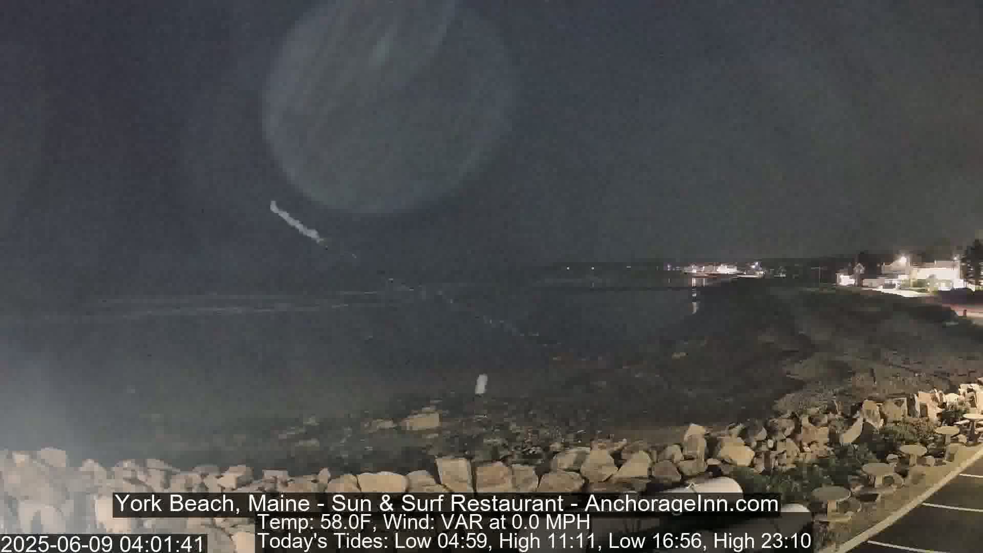A nighttime view of a rocky shoreline and calm dark ocean under a cloudy sky.