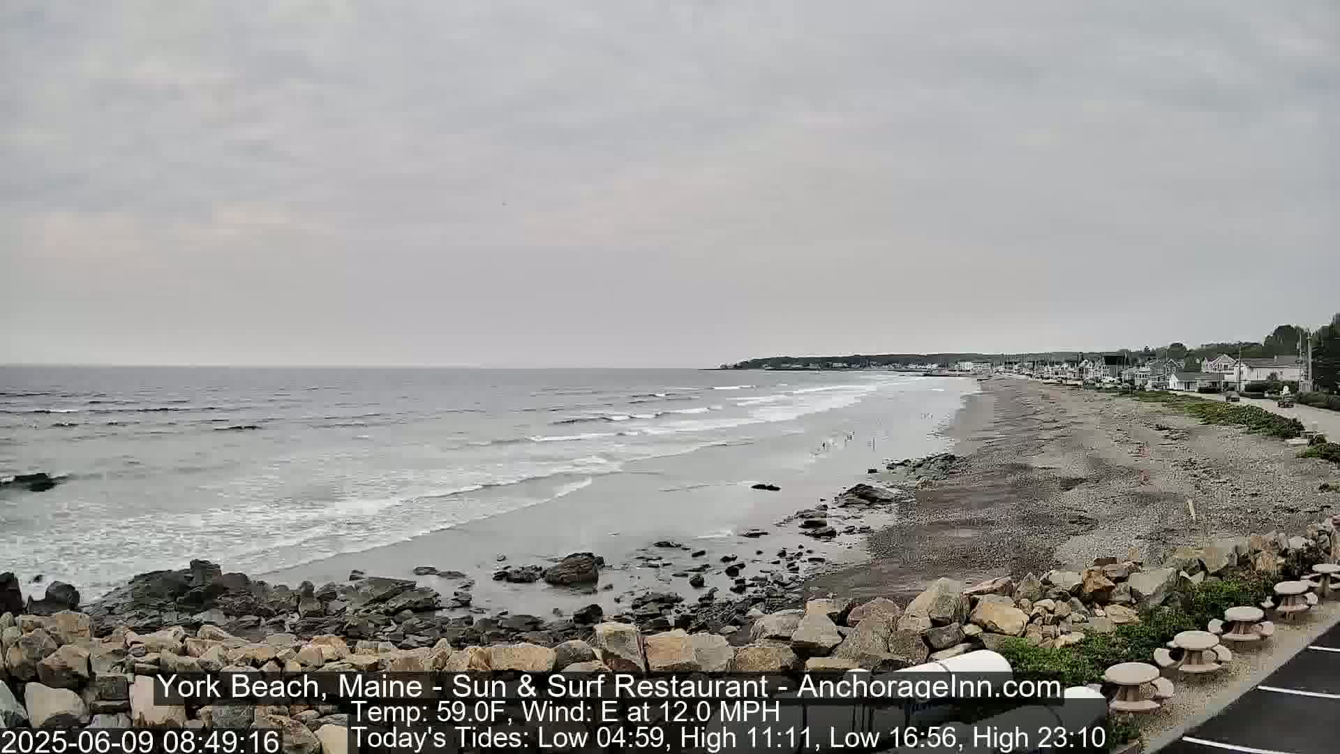 A rocky beach with a calm ocean under an overcast sky is shown, with a few buildings visible in the distance.