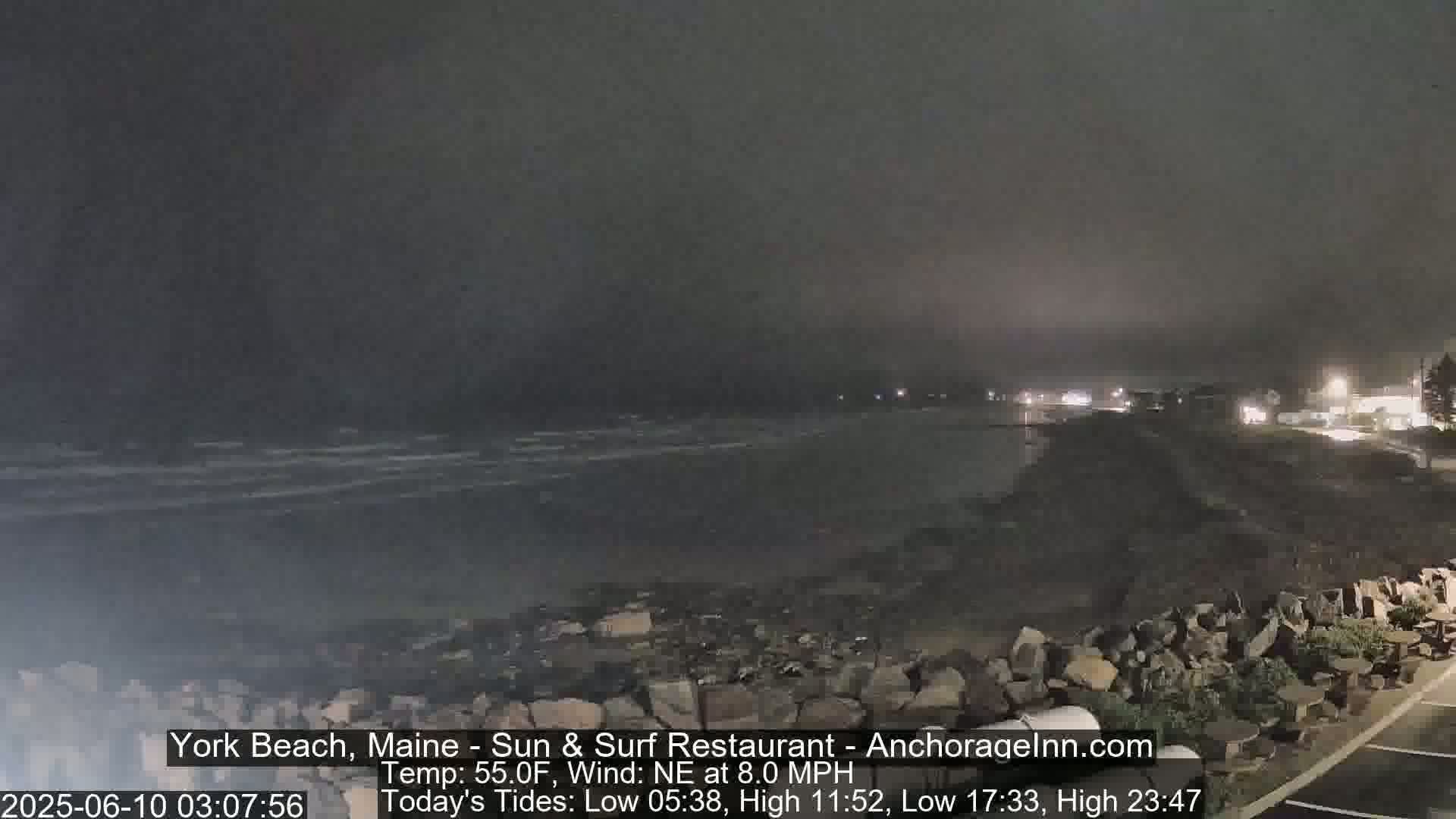 A nighttime coastal scene shows dark, choppy waves under a hazy sky, with a rocky shore and dimly lit buildings in the distance.