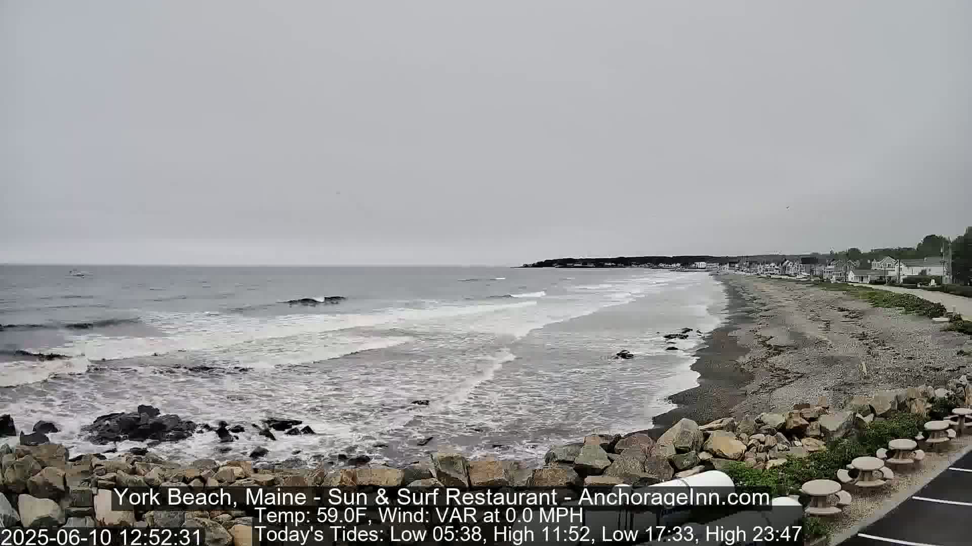 A gray, overcast day shows a beach with waves gently lapping the shore, backed by a line of houses and some picnic tables.