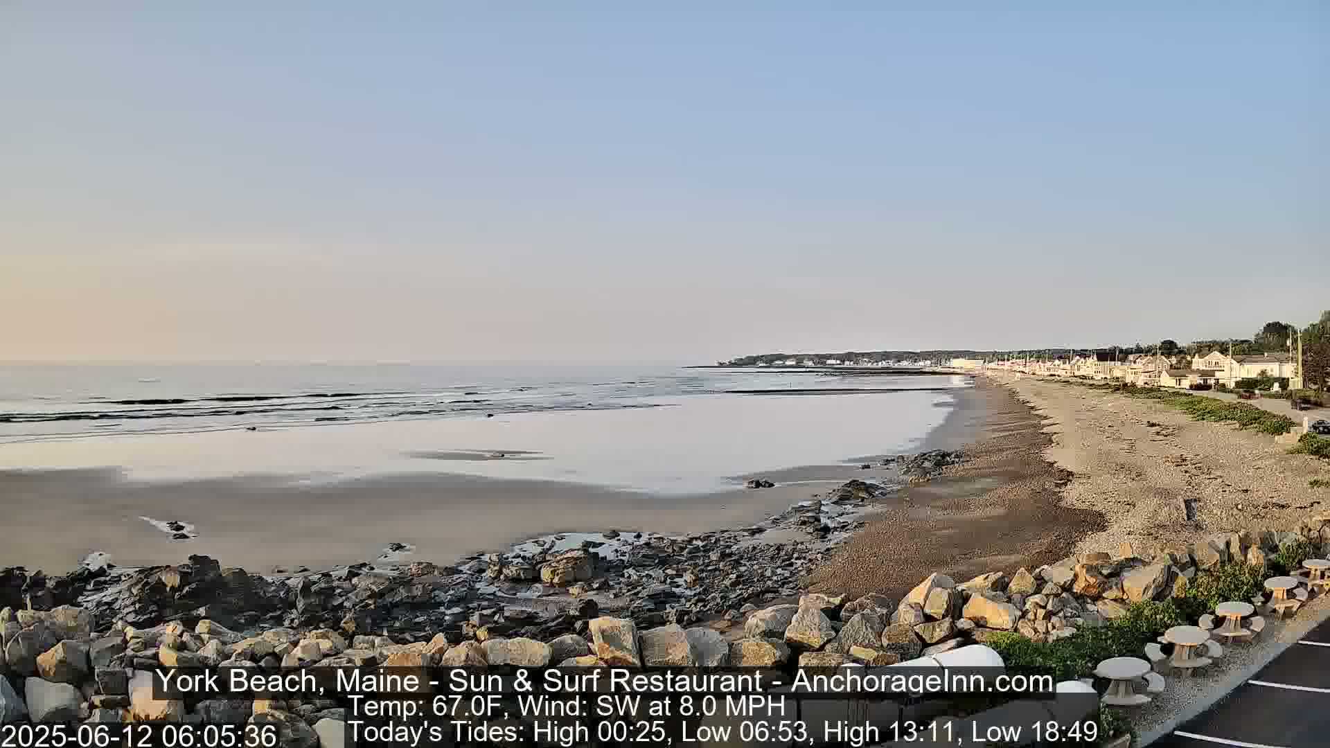 A sandy beach with rocky areas and calm ocean waves stretches toward a town under a clear, pale-blue sky.
