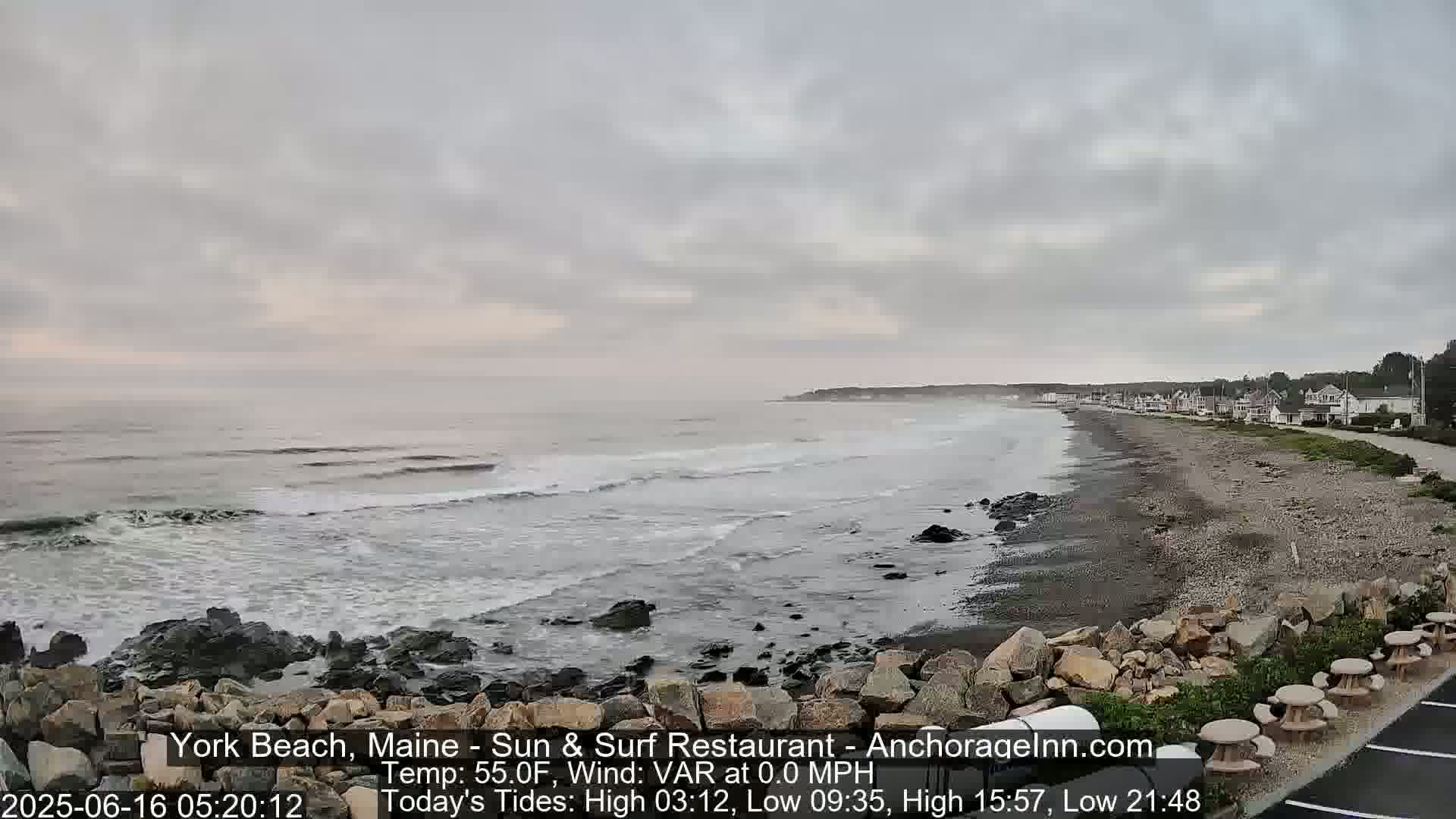 A gray, overcast day reveals a rocky shoreline with gentle waves lapping at the beach, backed by a row of houses.
