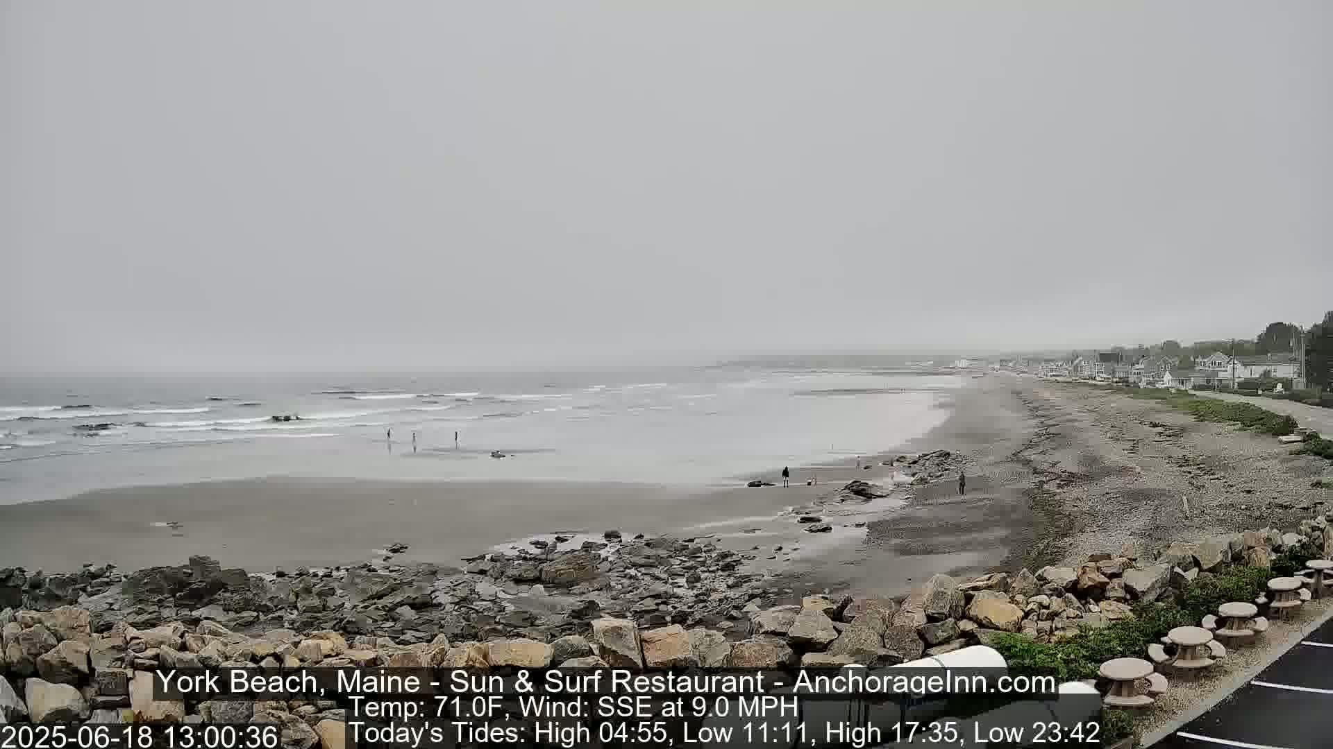 A hazy, overcast day at a beach shows a wide expanse of sand and rocky shoreline with a few people and small waves; a road and buildings are visible in the distance.