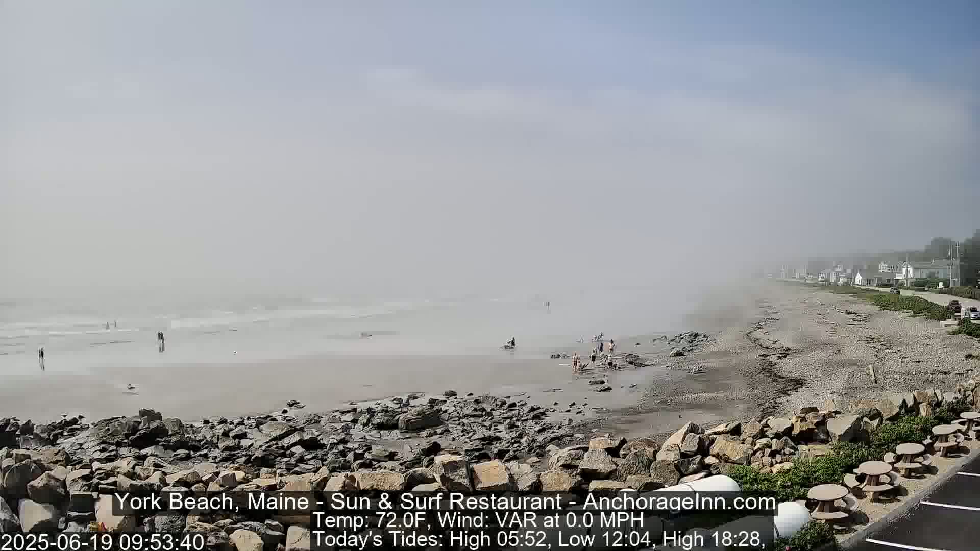 A foggy beach scene shows a rocky shoreline with a few people scattered across the wet sand, a calm ocean in the distance, and a road lined with buildings beyond.