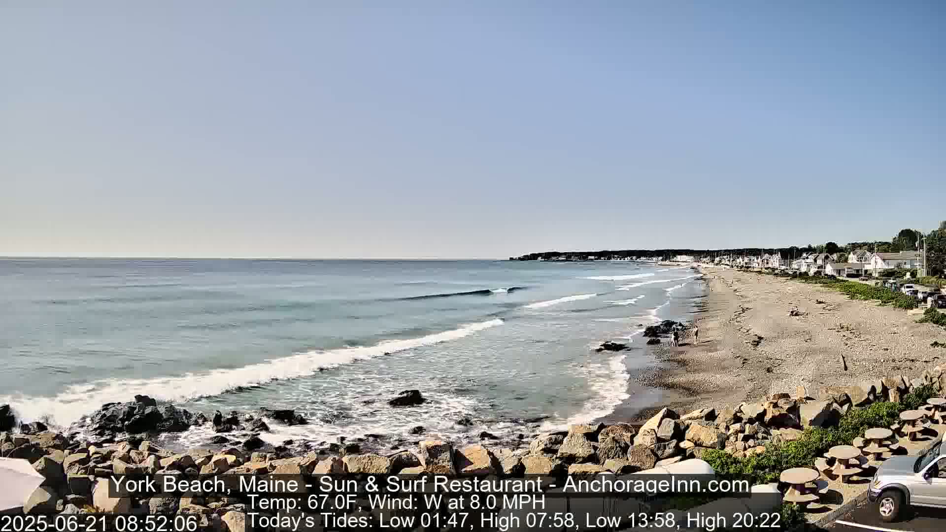 A sandy beach with gentle waves laps at a rocky shoreline under a clear sky; several houses are visible in the distance.
