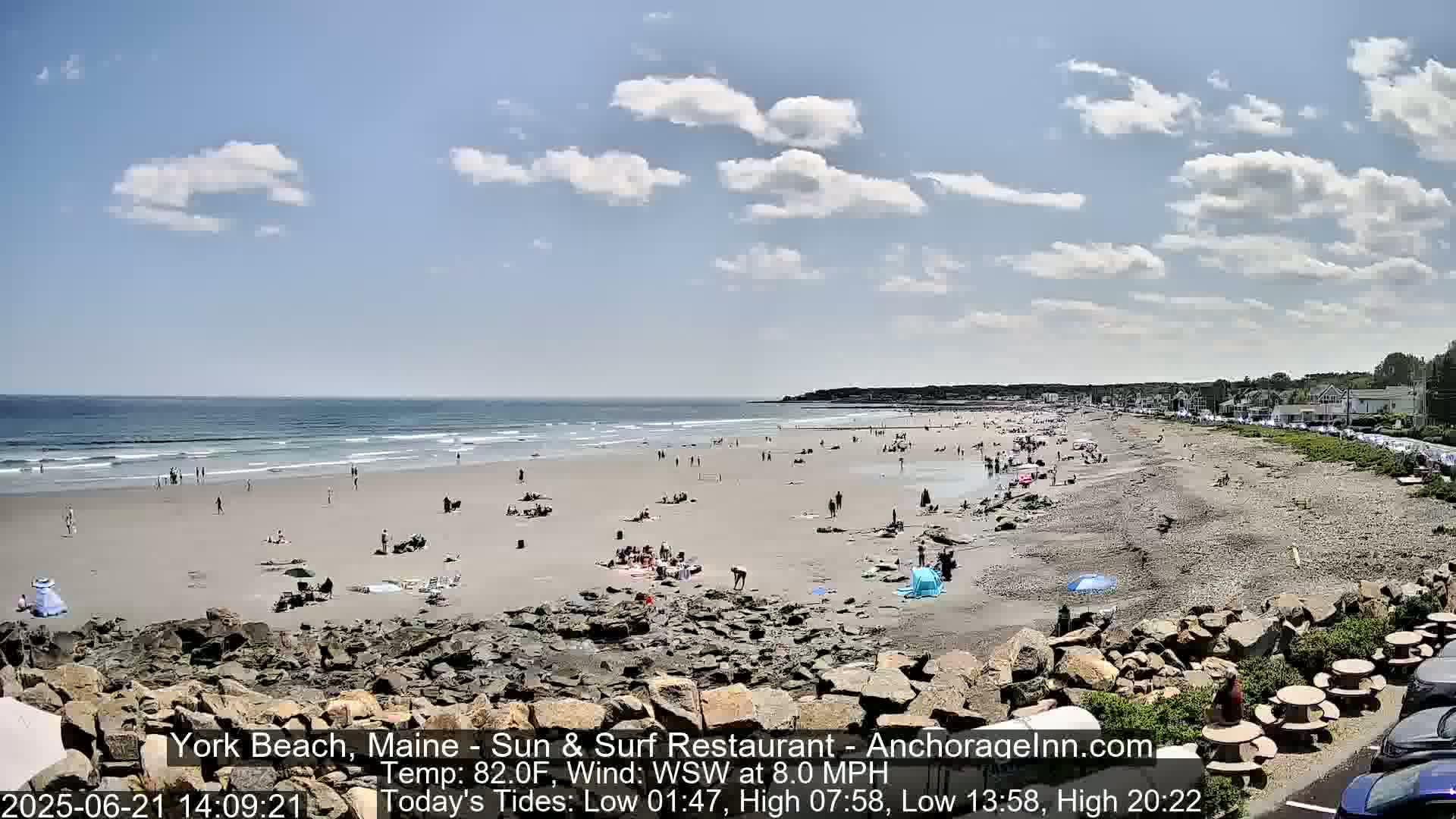 A sandy beach with many people on it and a partly cloudy sky is visible on a sunny day.