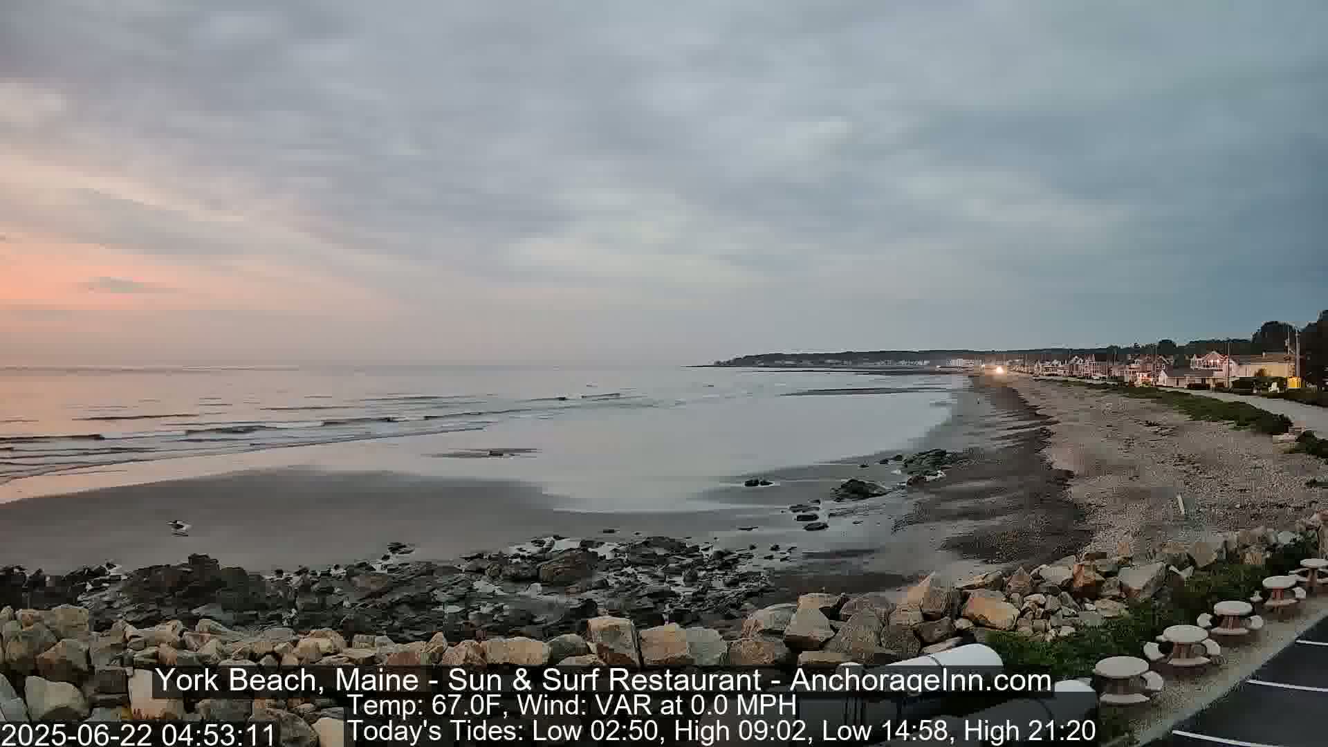 A mostly cloudy, pre-dawn scene shows a calm ocean receding from a rocky beach to reveal a wide expanse of wet sand, with a town visible in the distance.