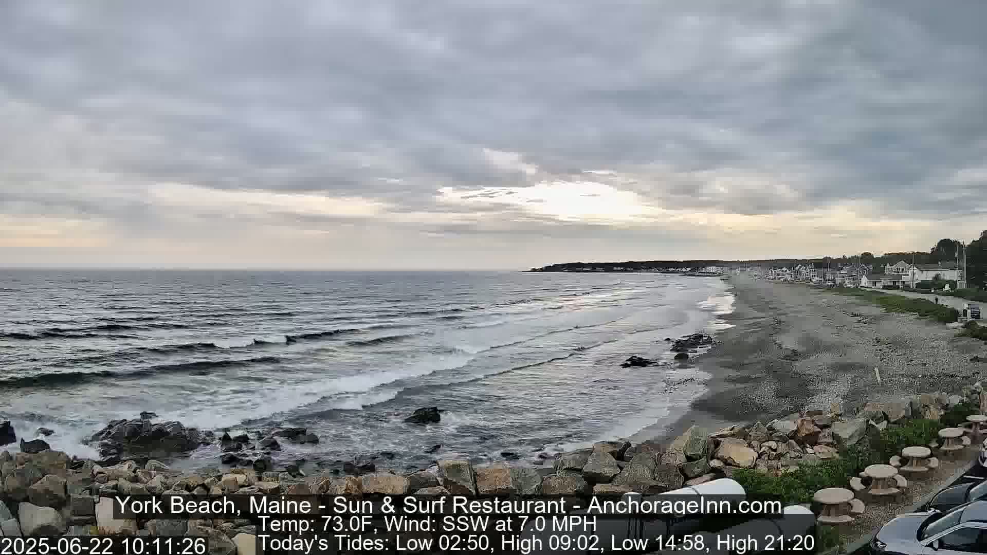 An overcast sky hangs over a rocky beach with gentle waves lapping the shore, and a town visible in the distance.
