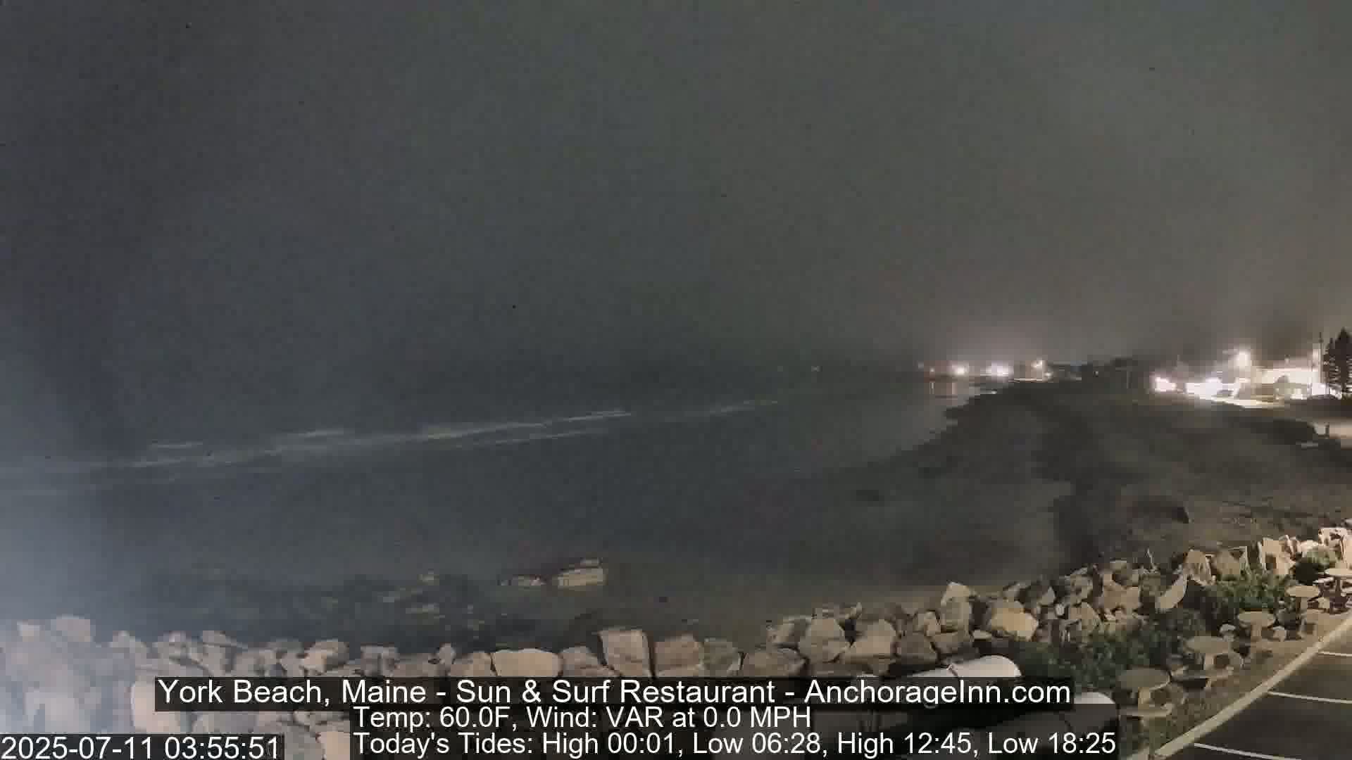 A dark, nighttime beach scene with calm water and a rocky shore under hazy conditions.