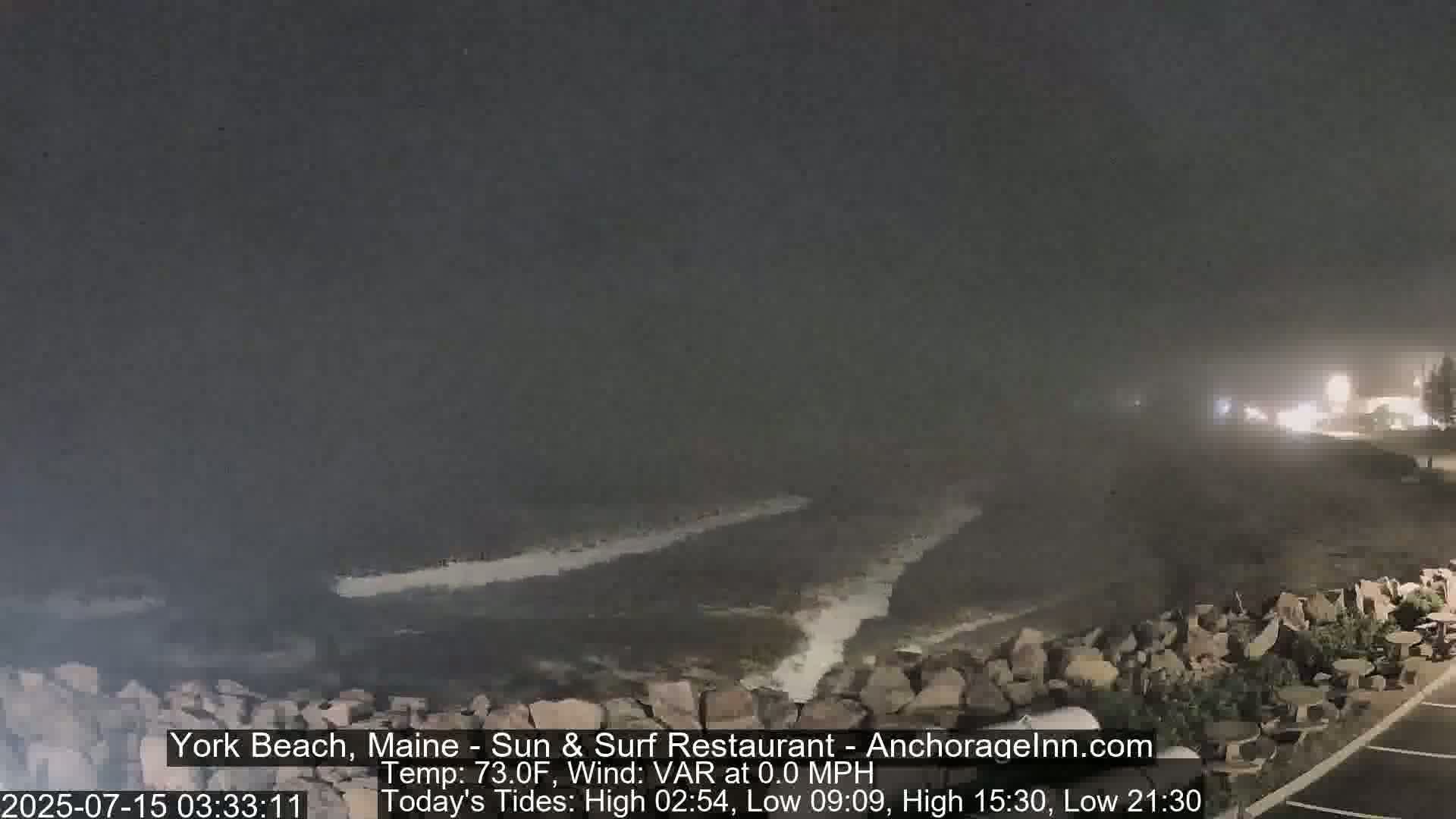 A dark, nighttime scene shows a rocky shoreline with waves crashing against it under a hazy, dark sky.
