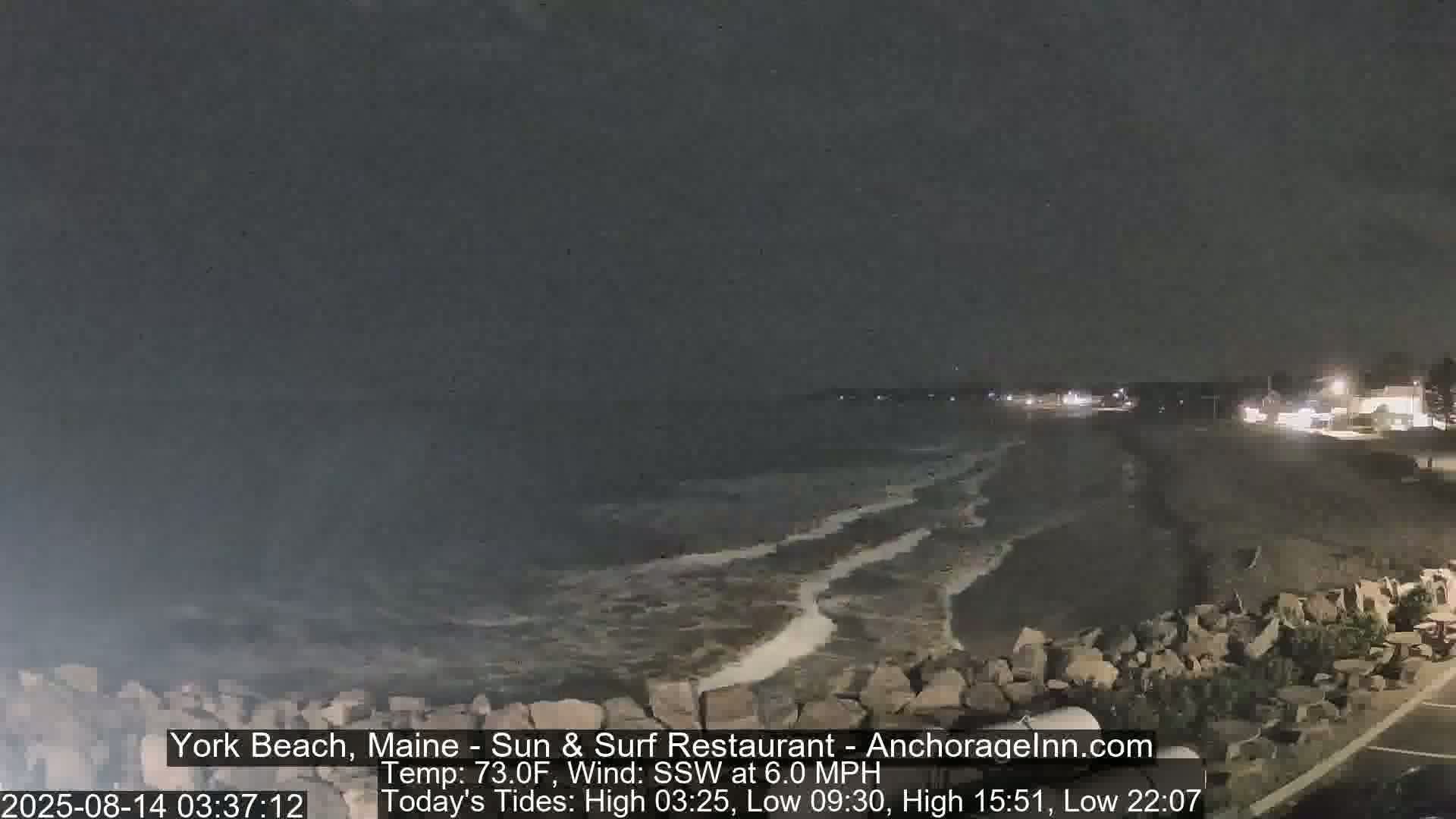 A dark, nighttime beach scene shows gentle waves lapping the shore under a mostly clear sky.