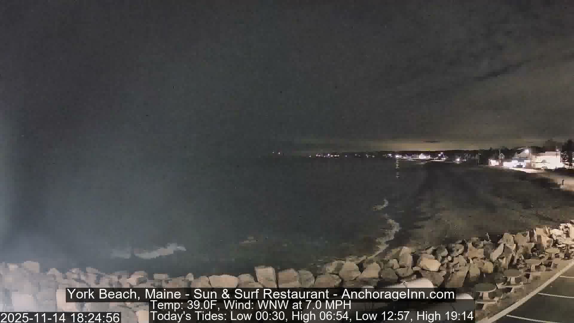 A nighttime view under a cloudy sky reveals waves breaking on a rocky and sandy beach, with distant town lights illuminating the coastline and some outdoor seating in the foreground.