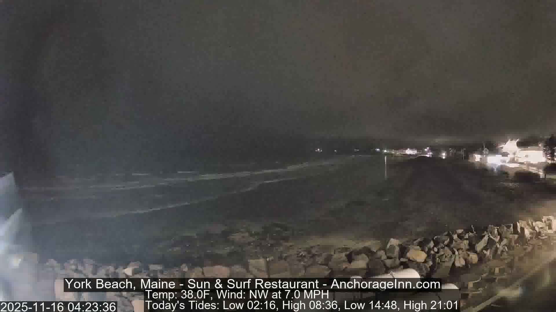 A dark, cloudy night reveals waves breaking on a beach with a rocky foreground and distant illuminated buildings along the coastline.